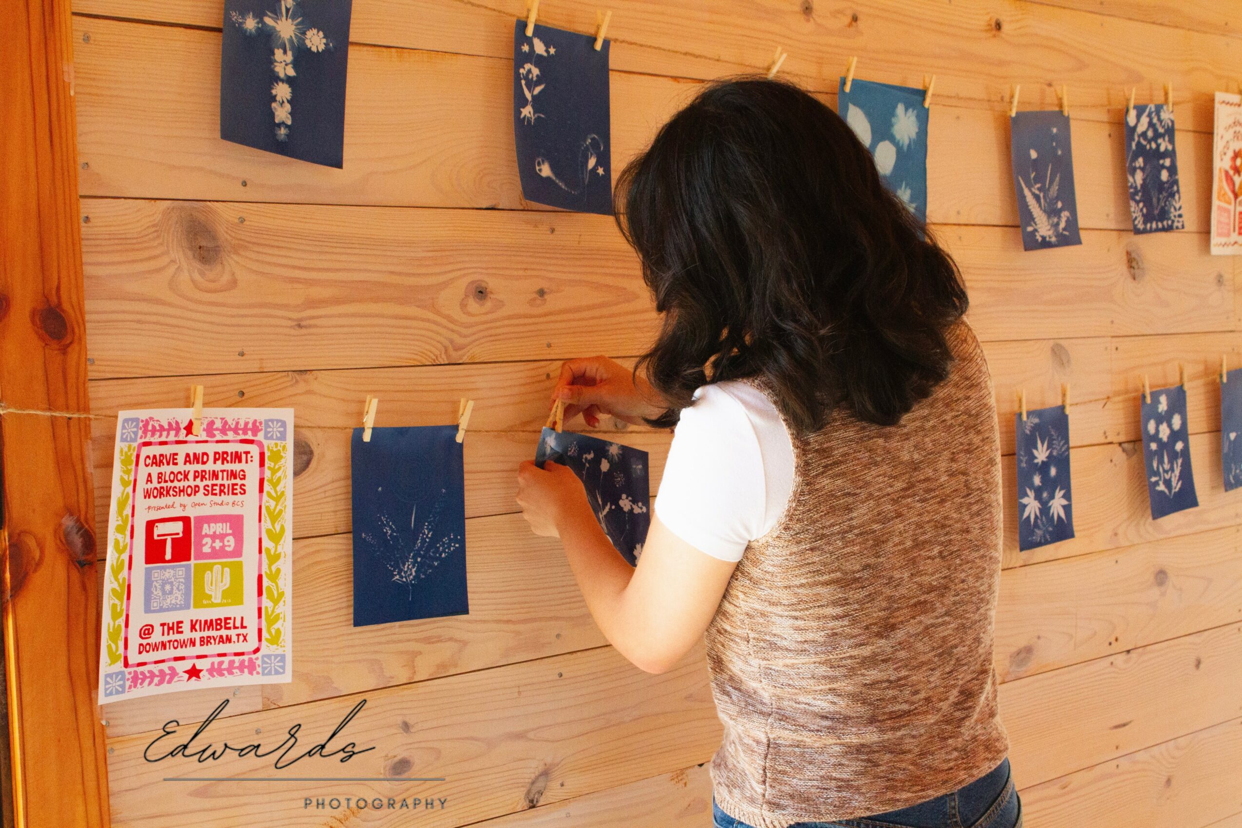 Child with dark hair, wearing a white shirt and brown vest, looks at hanging artwork on wooden wall.