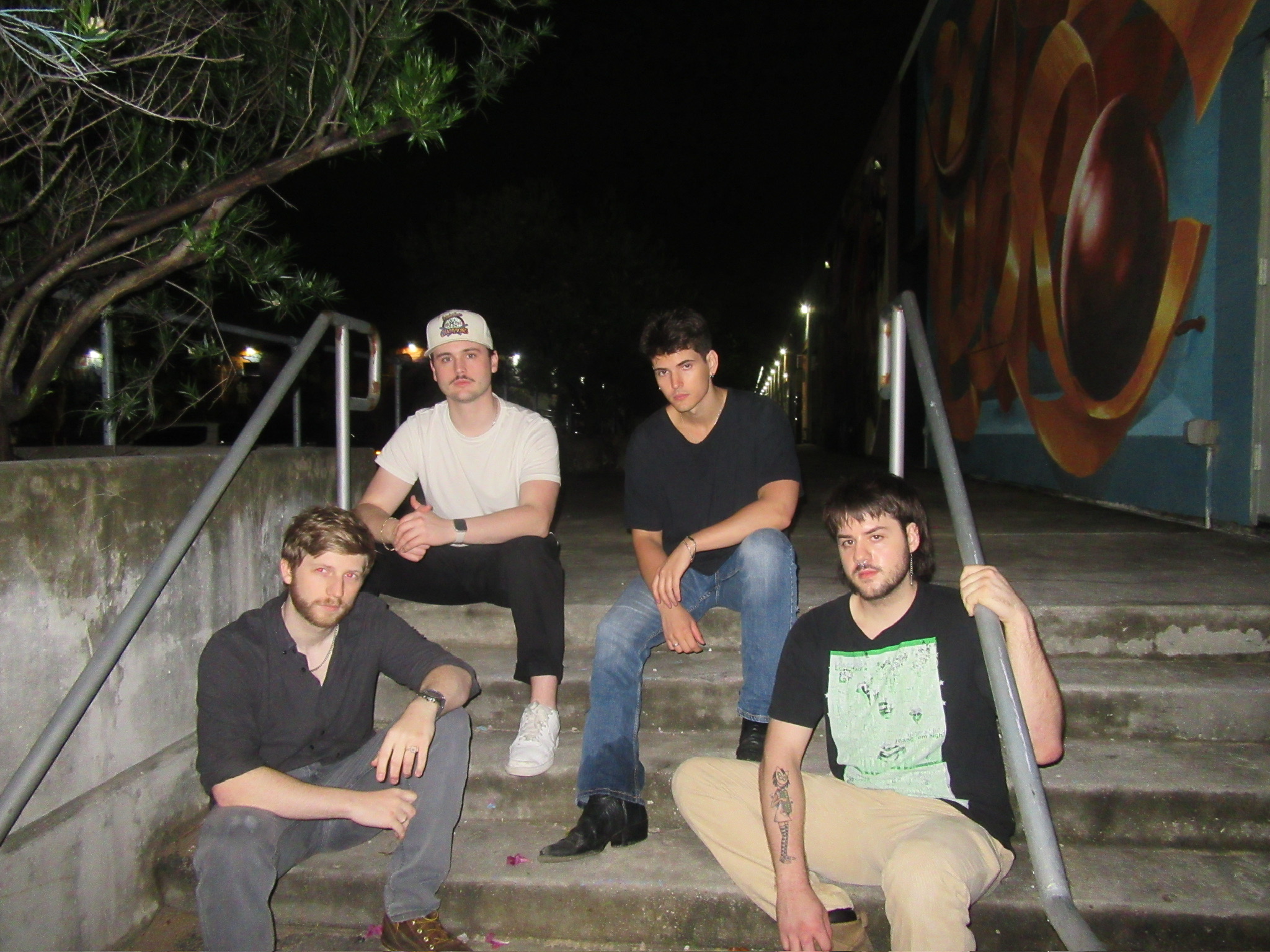 Four young men sitting on stairs outdoors at night, some leaning on railings, with trees and a mural in the background.