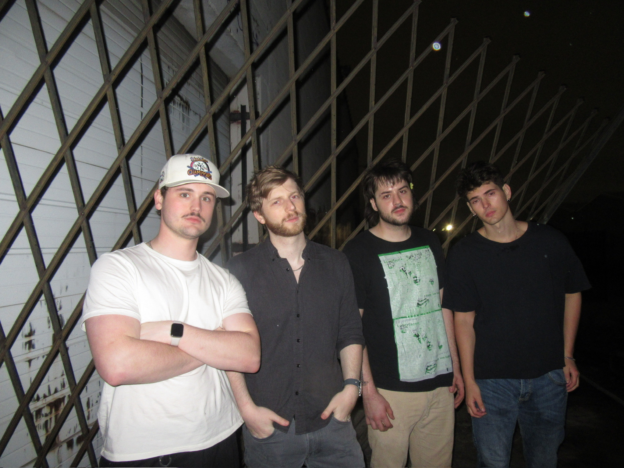 Four young men standing outdoors at night, with a geometric metal structure behind them.