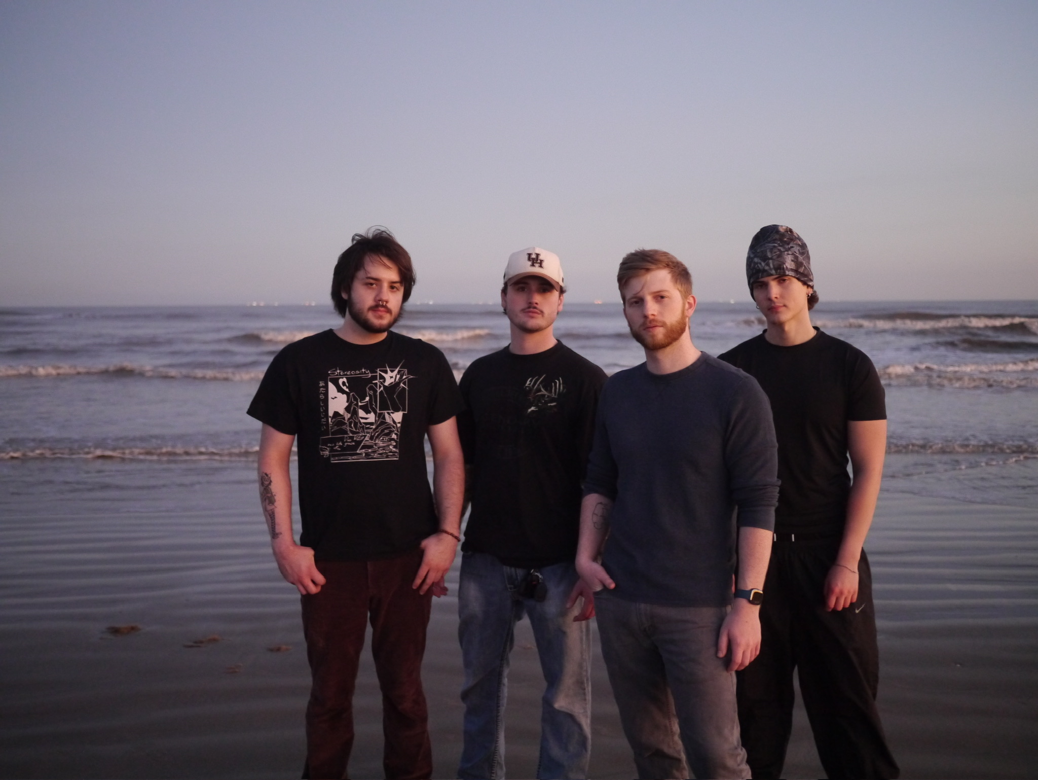 Four men stand on a beach with ocean waves in the background, facing the camera, during sunset or sunrise.
