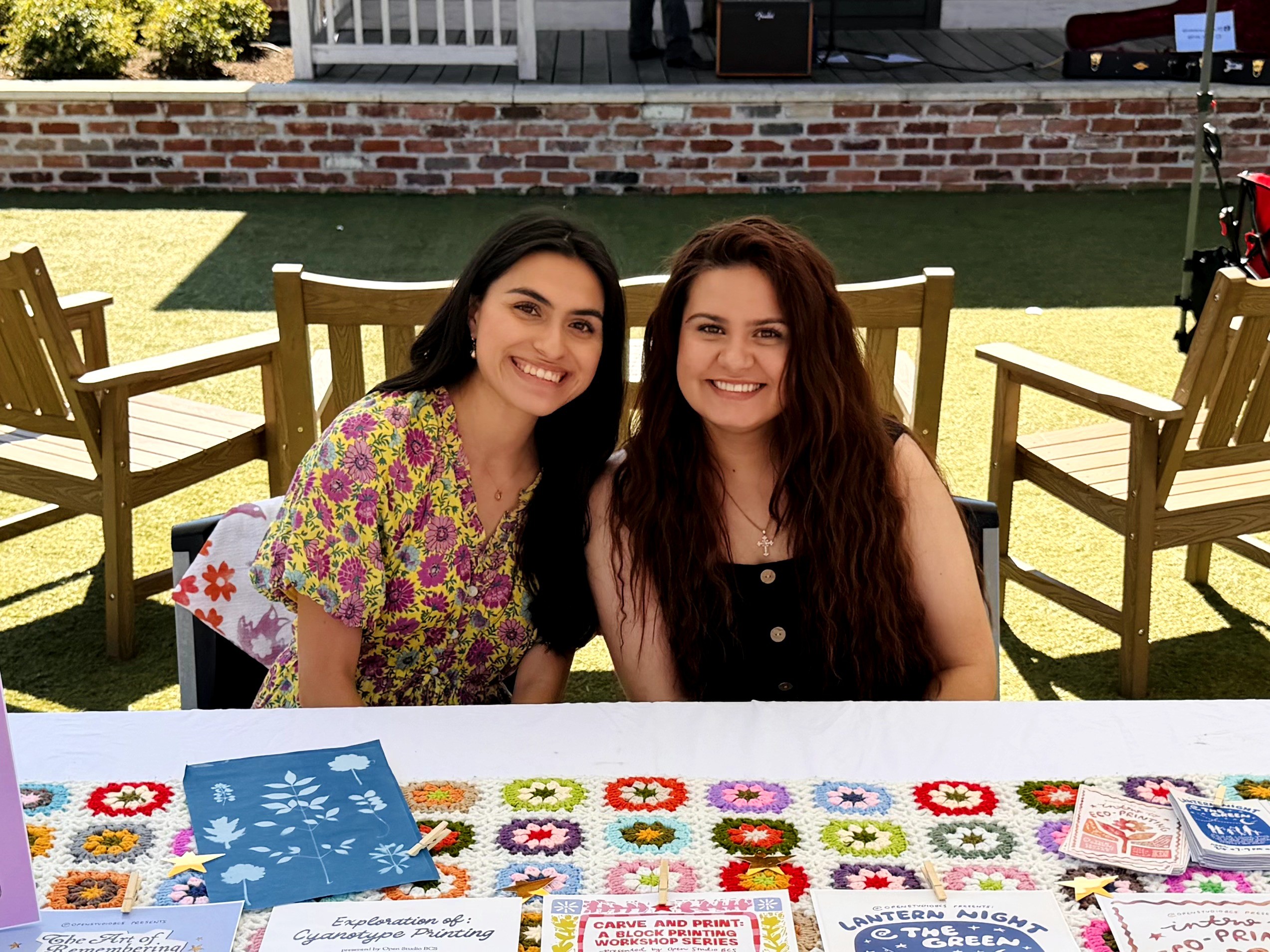 Two women sit at a table outdoors, smiling, with chairs and a grassy area in the background.