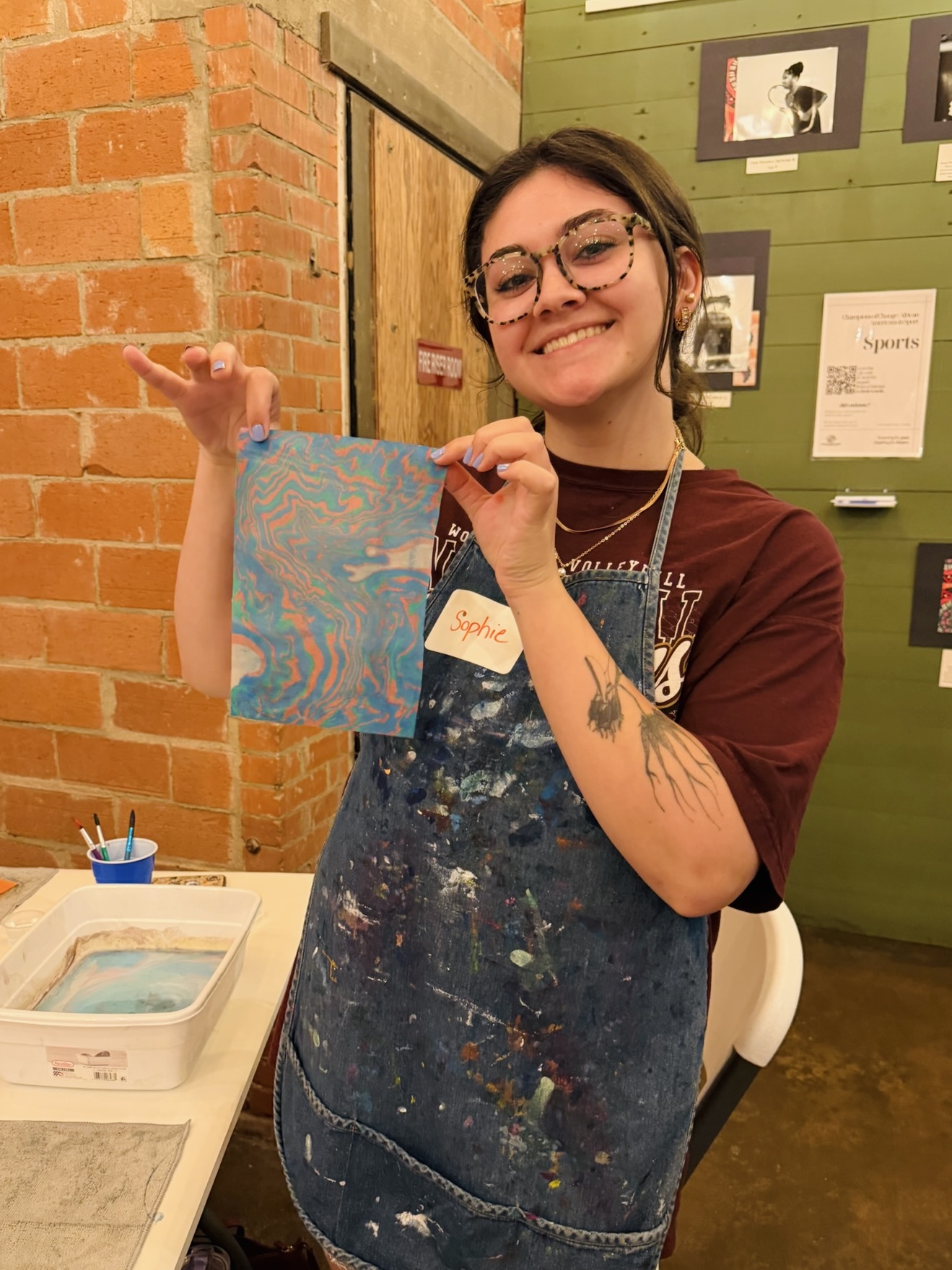 Young woman with glasses smiling, holding a colorful abstract painting in a creative space.