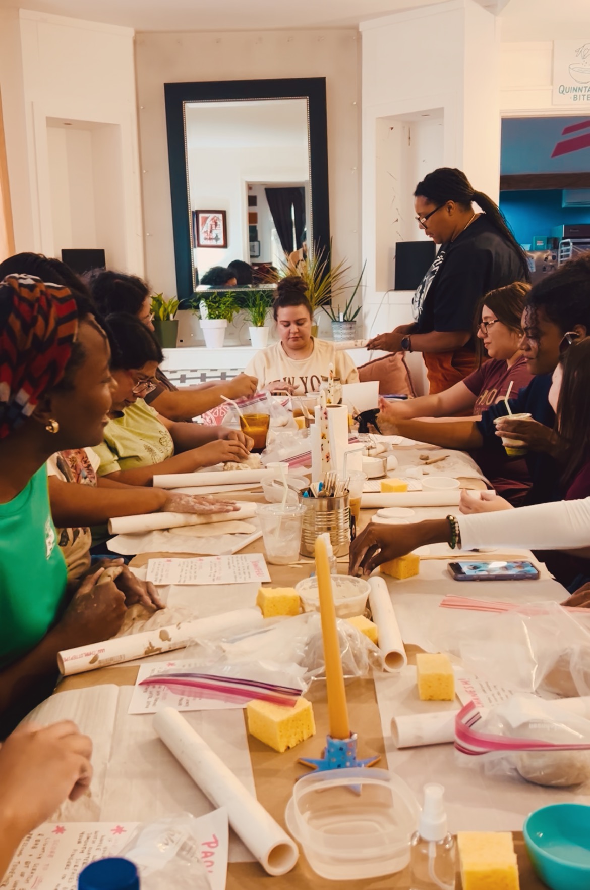 Group of people sitting around a table with papers, drinks, and yellow candles in a room with a mirror and plants.