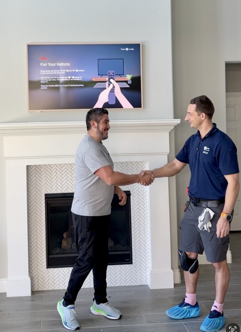 Two men shake hands in front of a fireplace and television, one wearing a gray shirt and the other in a blue uniform, in a living room.