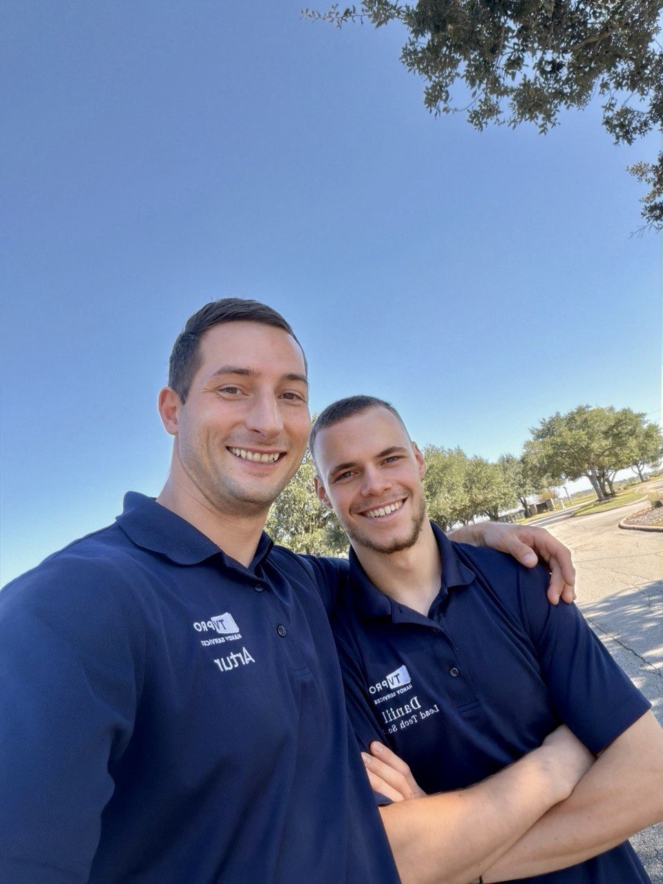 Two men in navy blue shirts smiling outdoors, one with arm around the other, under a clear blue sky.