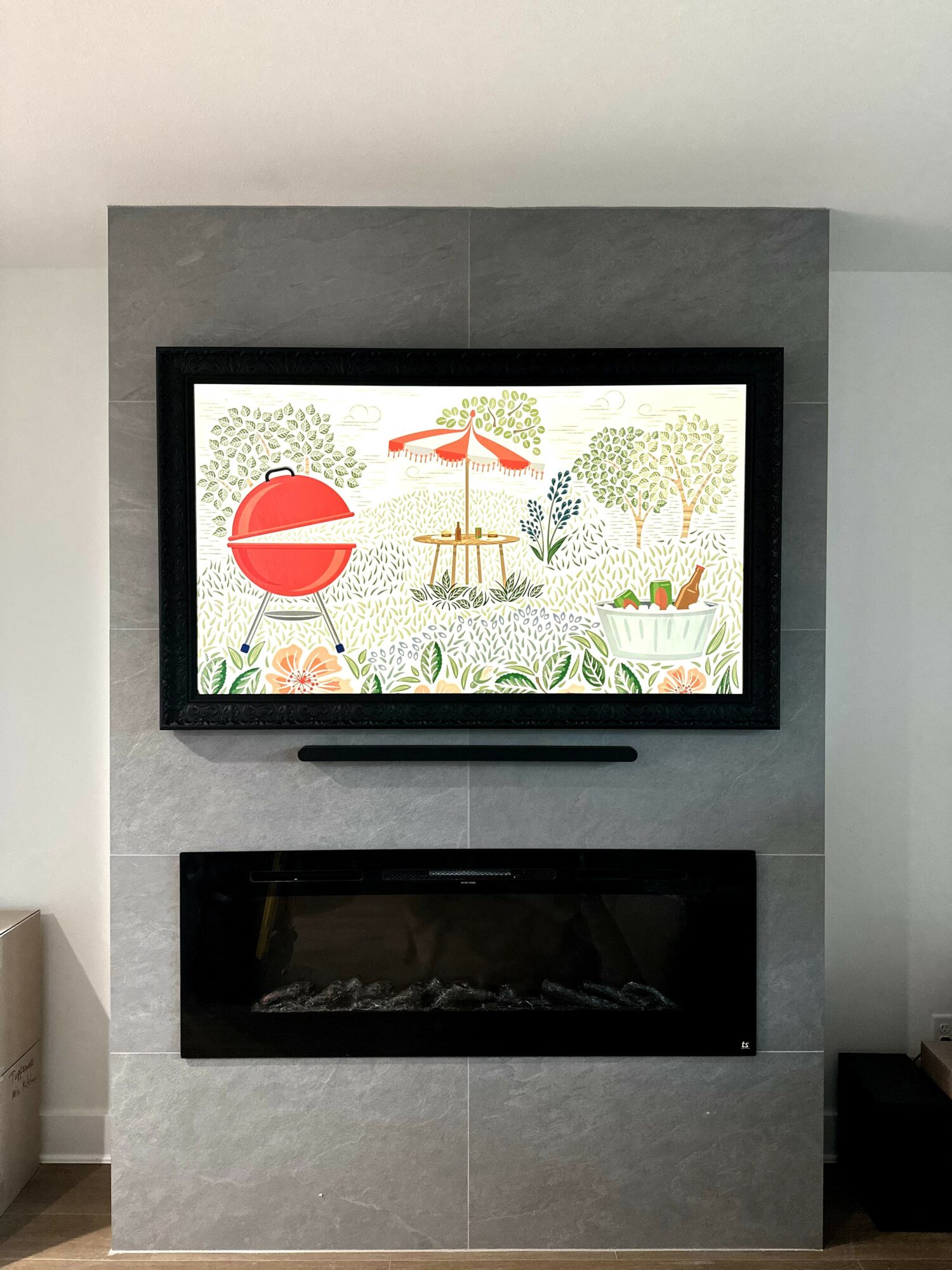Framed colorful illustration of trees, a bench, and a large red circle, above a modern fireplace, on a gray wall.