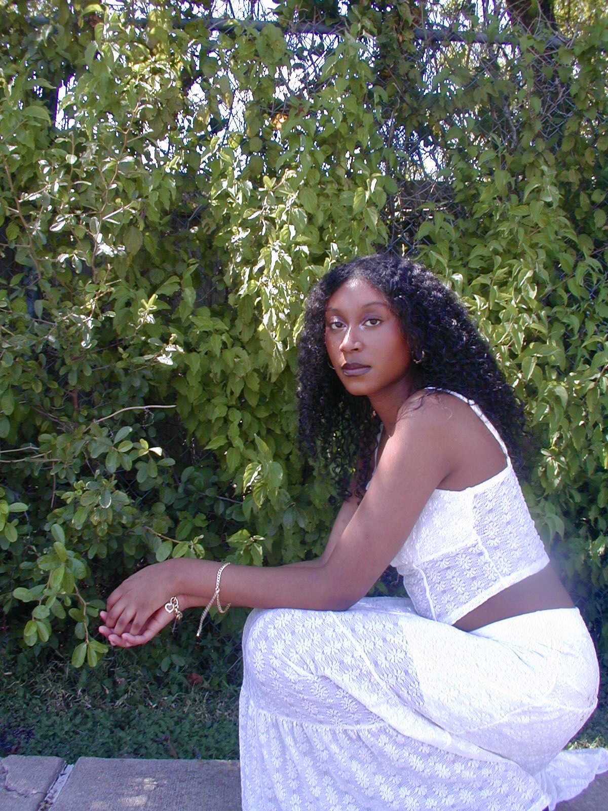 Young woman with curly hair sitting outdoors in front of green foliage, wearing a white sleeveless top and pants.