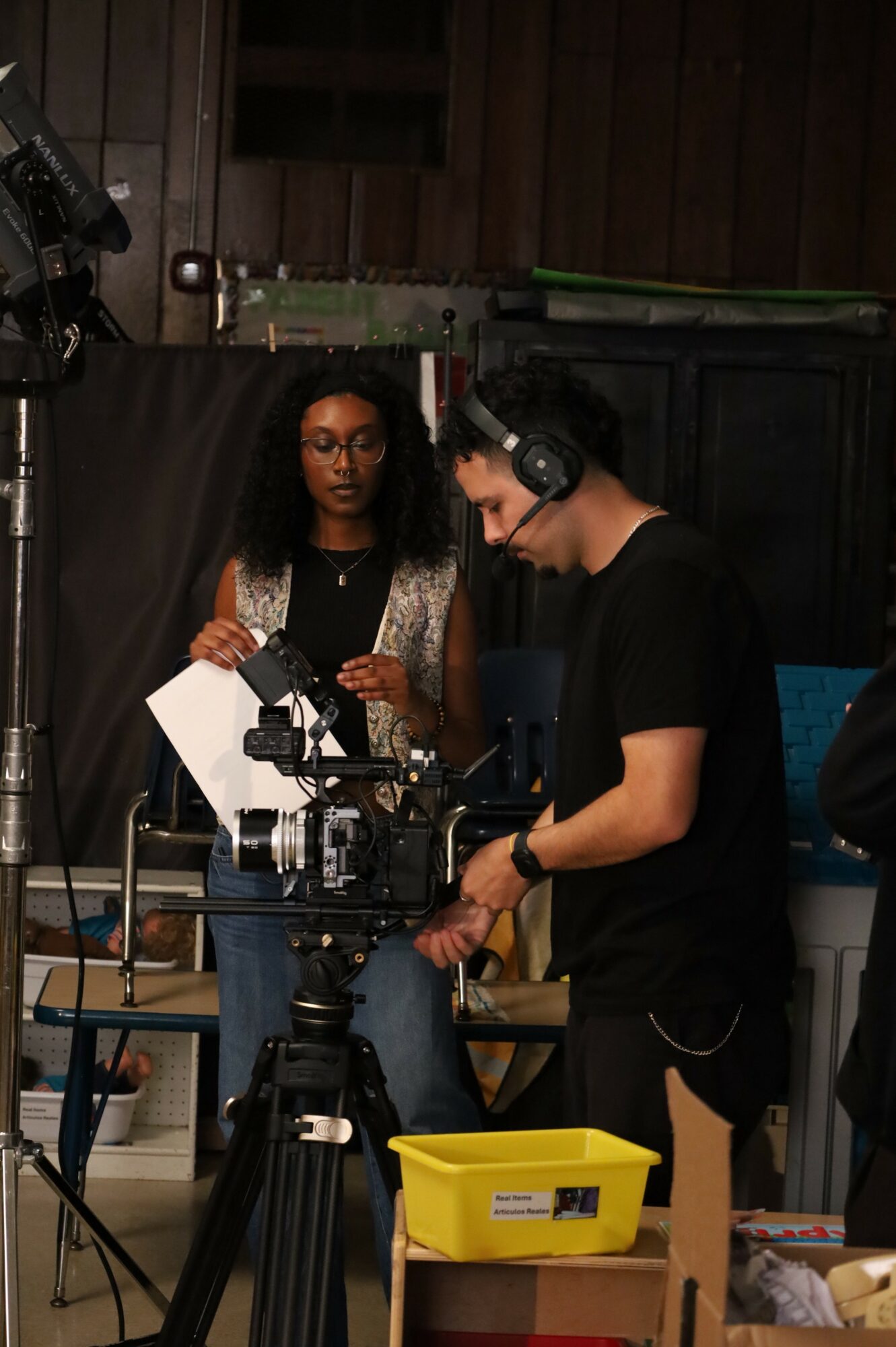Two people working with camera equipment in a studio, one woman holding papers and one man adjusting the camera, yellow box nearby.