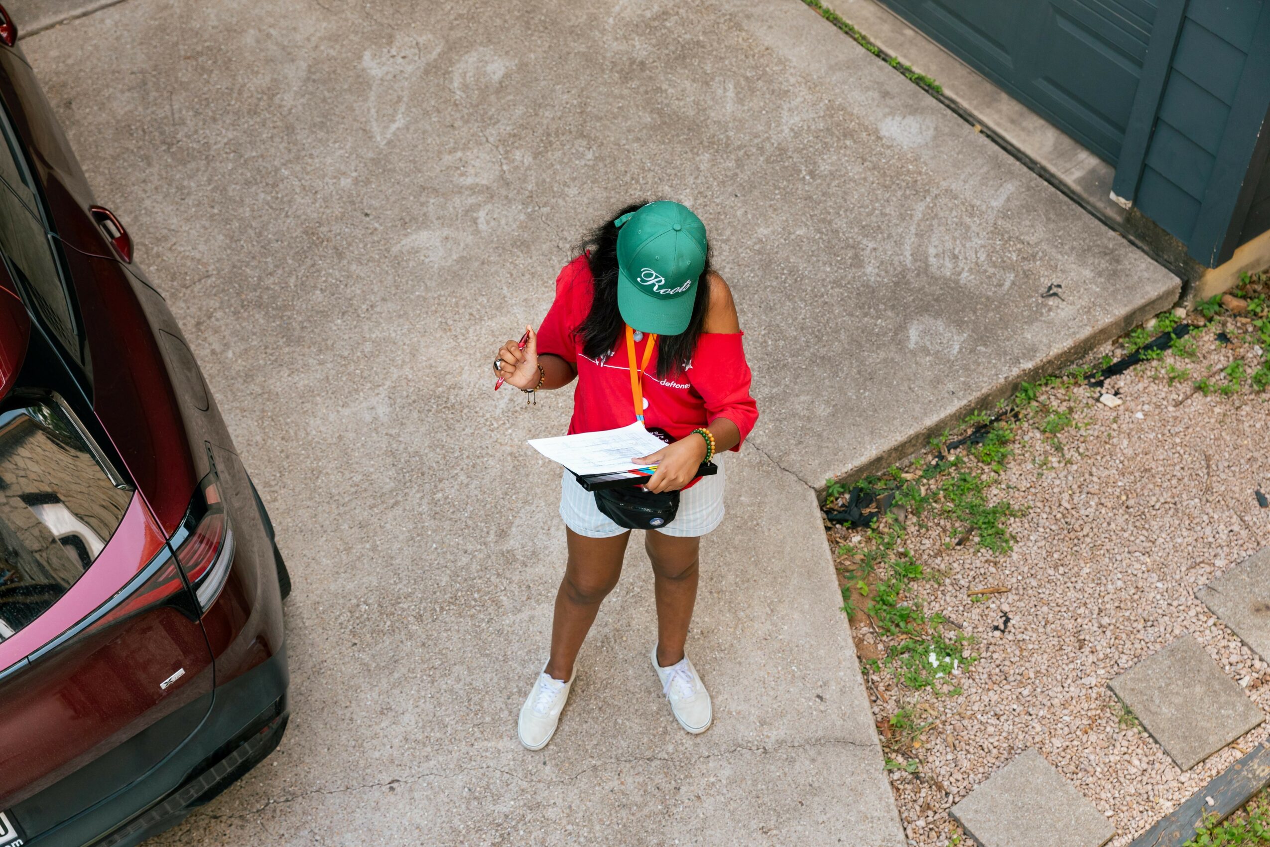 Person wearing a green cap, red jacket, and white shoes holding a paper, standing on concrete near a car and a blue fence.