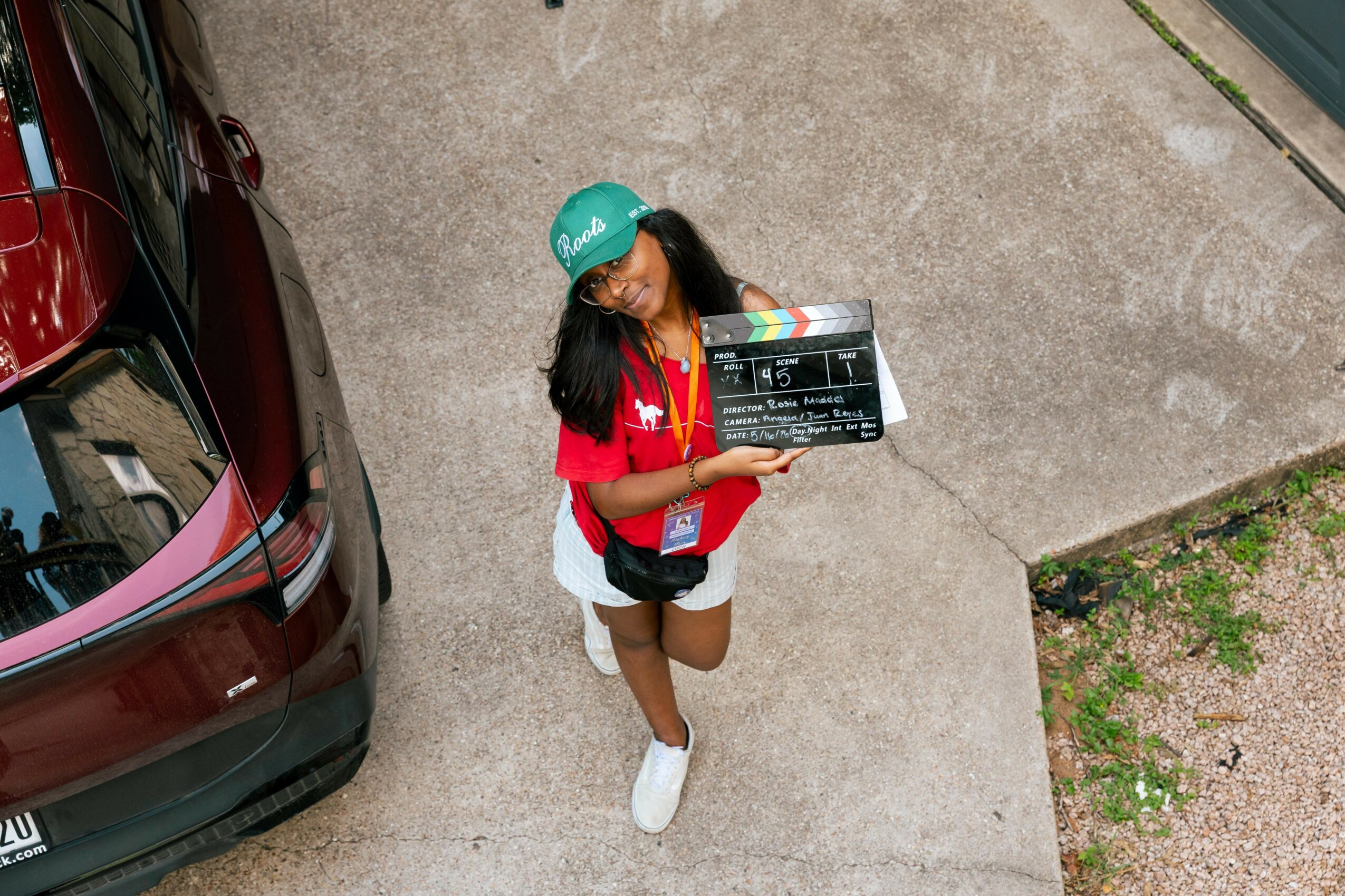 A woman wearing a teal helmet and red shirt stands on a concrete surface holding a clapperboard, next to a parked car.