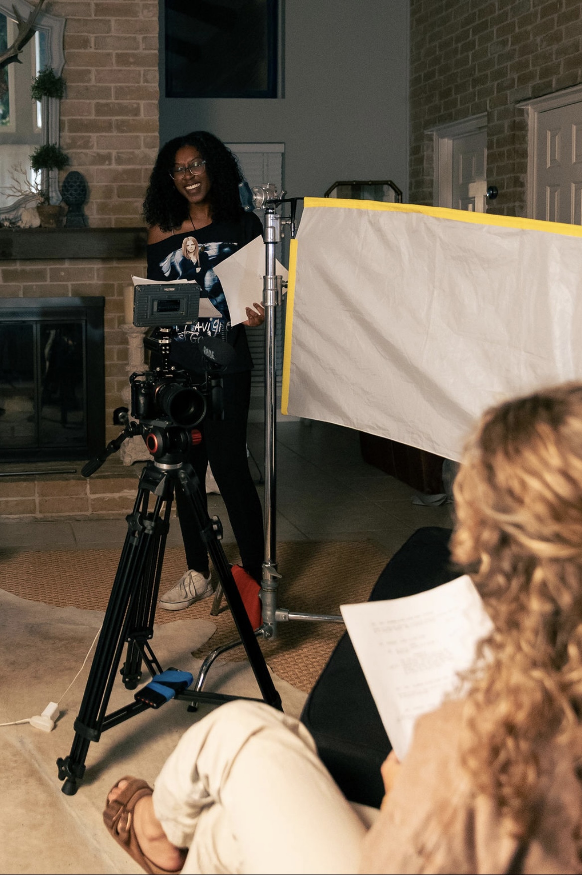 Woman with curly hair and glasses stands behind camera, holding papers, in a room with brick wall and lighting equipment.