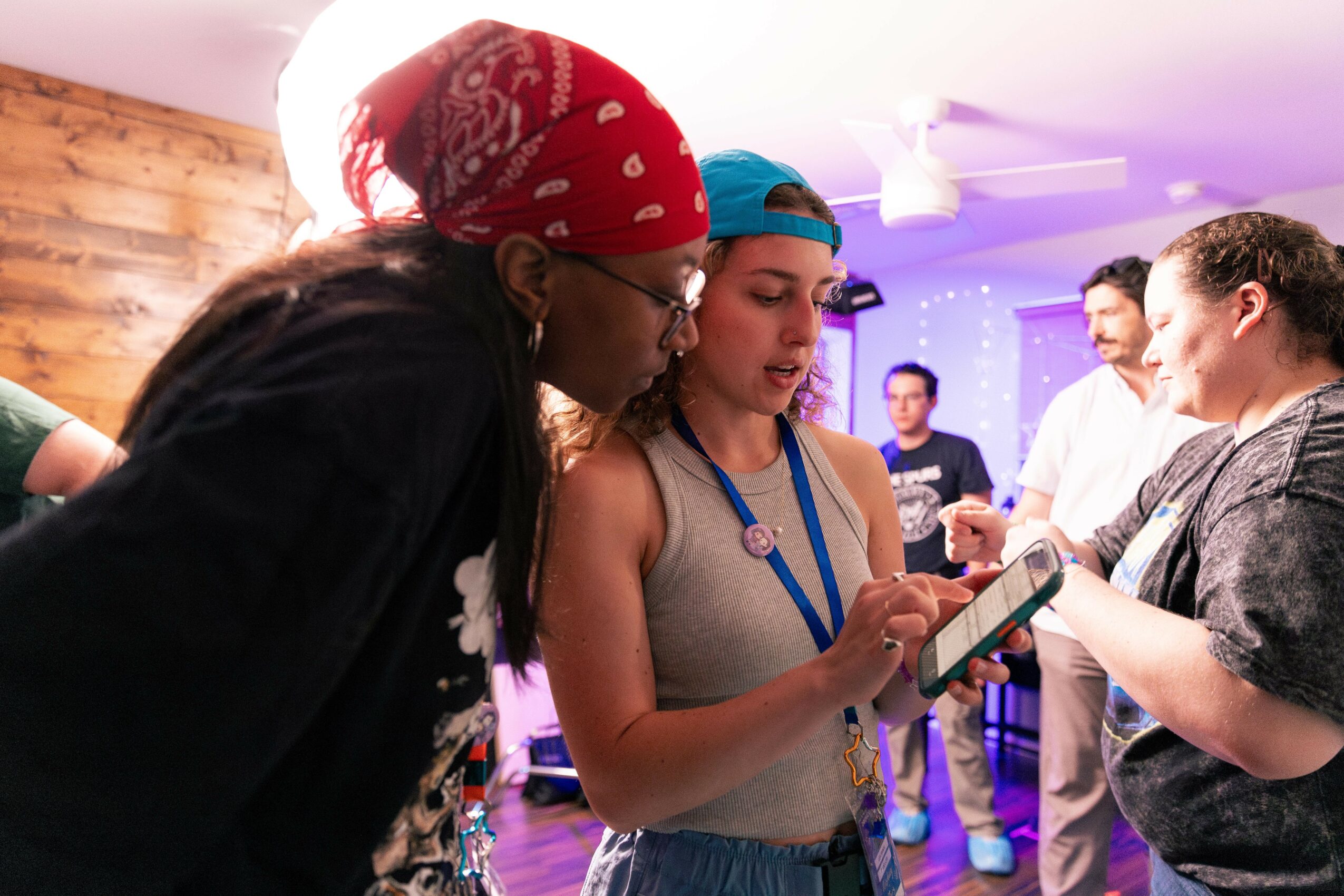 Three women are engaged in a conversation indoors, with other people in the background, some holding papers or devices.
