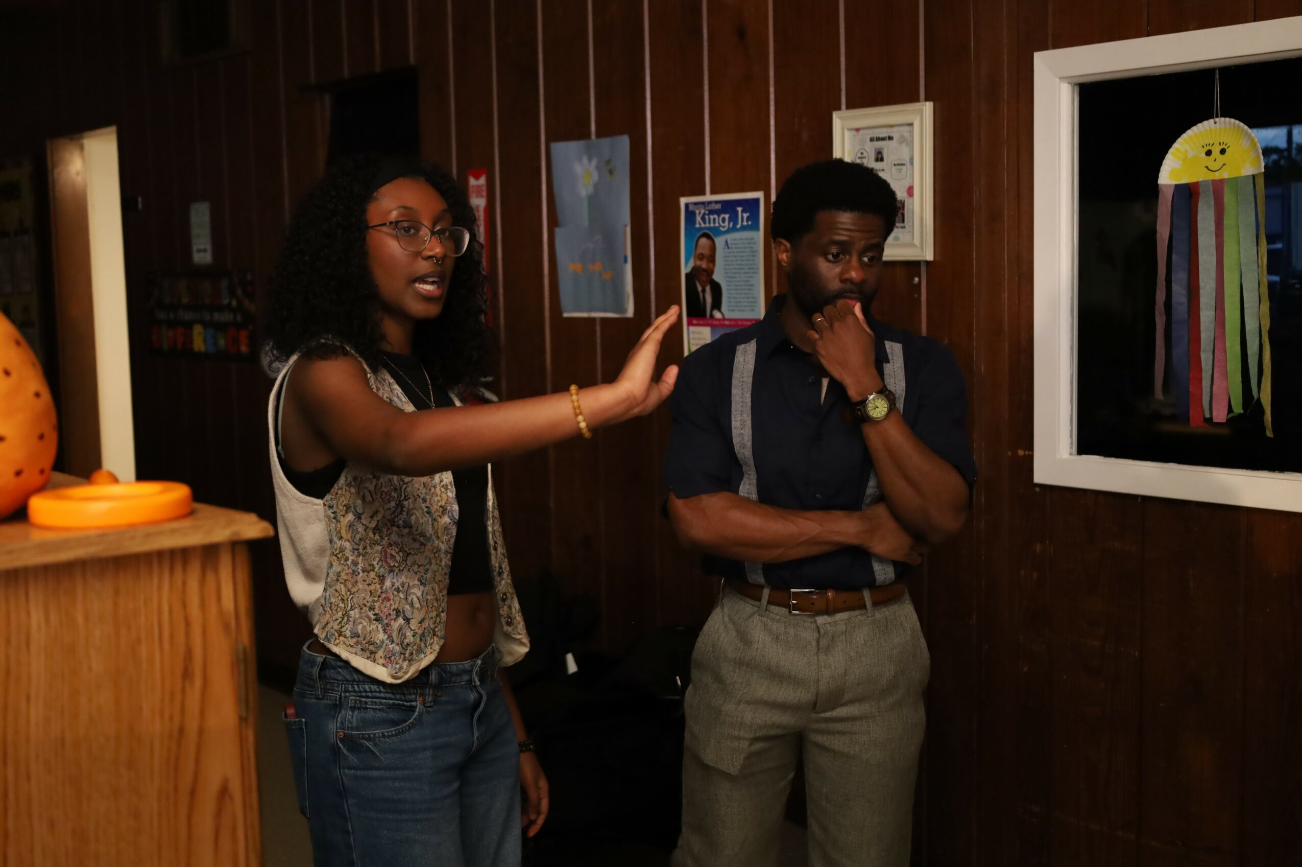 Two people stand in front of a wall with framed pictures, observing a colorful artwork on the wall.