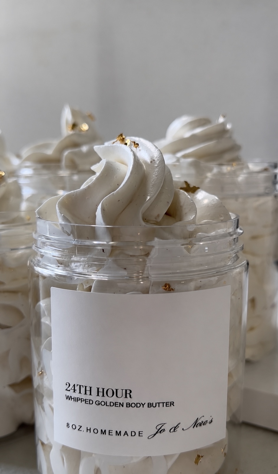 Cream-colored whipped body butter in small jars with white labels, topped with gold flakes, on a light background.