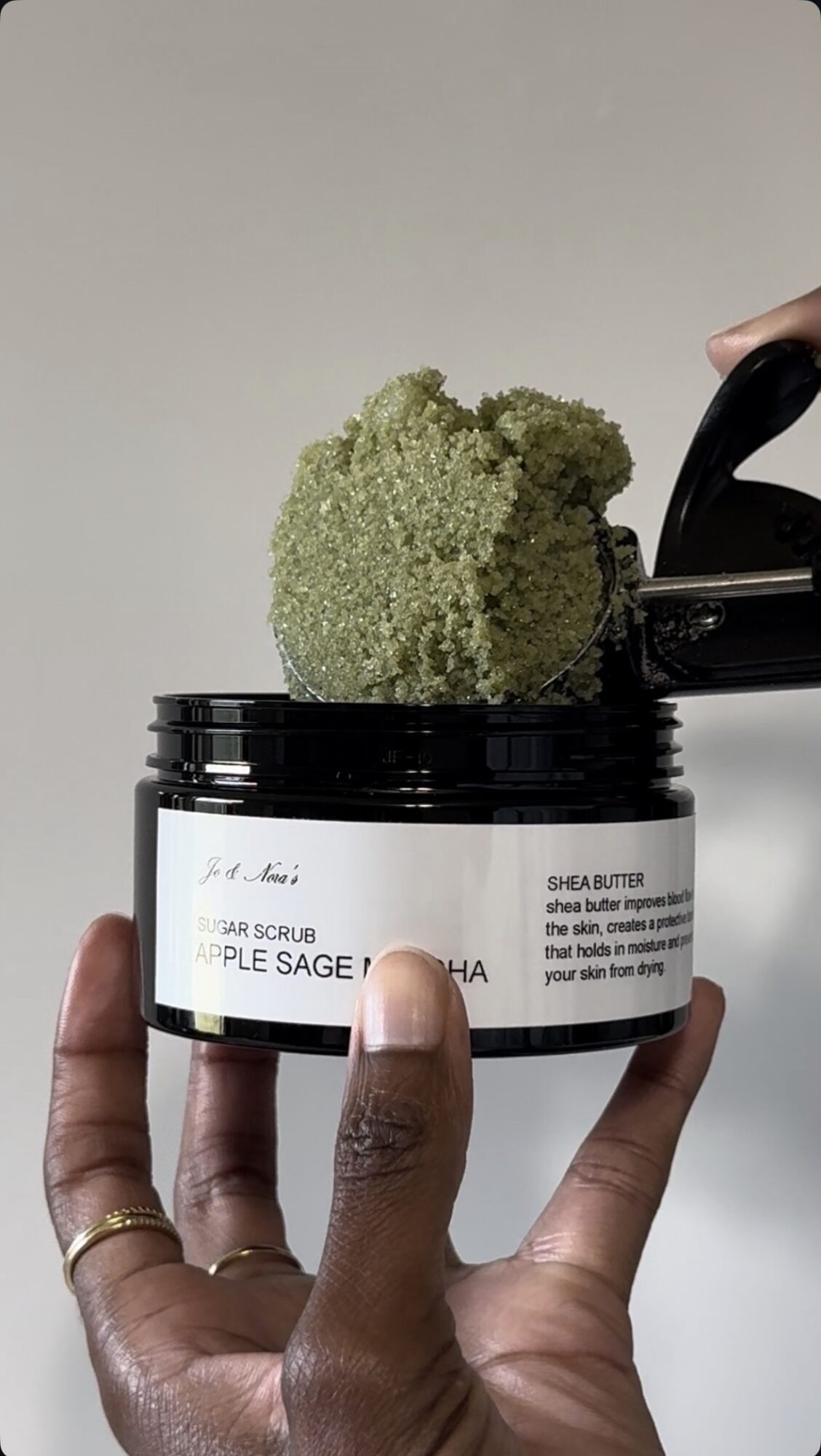 Hand holding a jar of skincare product with a green exfoliating scrub and a black spatula.