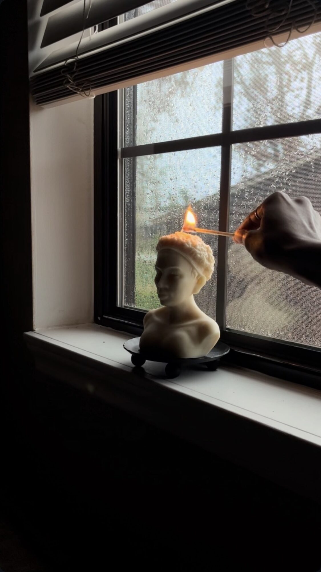 Person lighting a candle on a bust sculpture placed on a windowsill, with a window showing trees outside.
