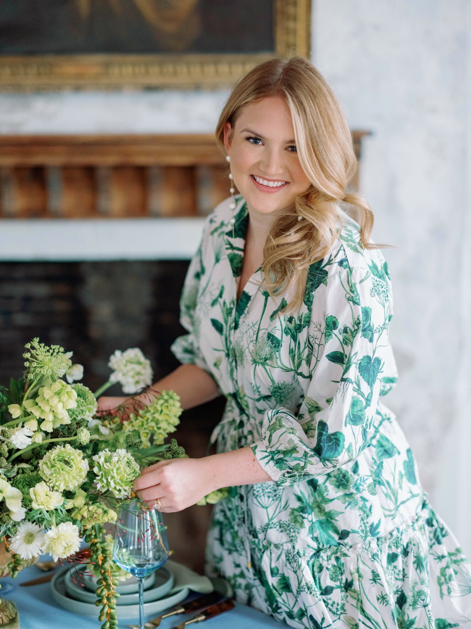 Smiling woman with blonde hair in a floral dress sitting at a table with a flower arrangement, fireplace, and painting in background.