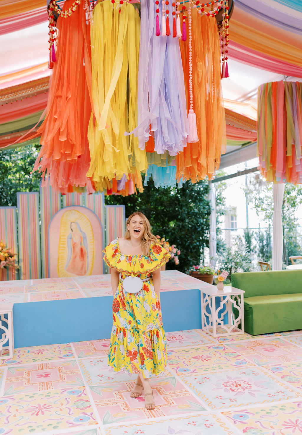 Person in colorful dress standing under hanging decorations, smiling, in decorated indoor space with rainbow-themed background.