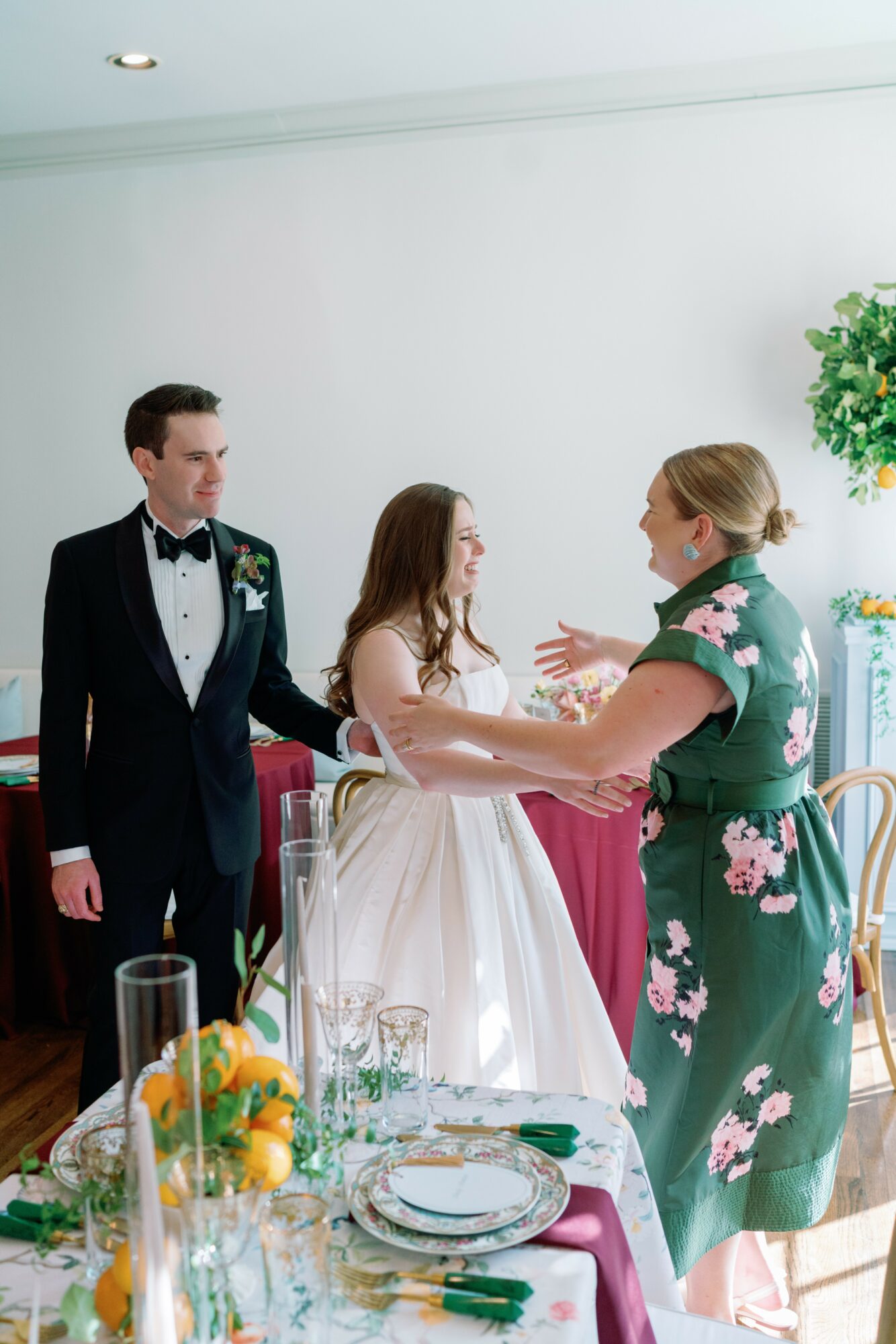 Three people, a bride in a white dress, a groom in a tuxedo, and a woman in a floral dress, are indoors, holding hands and talking.
