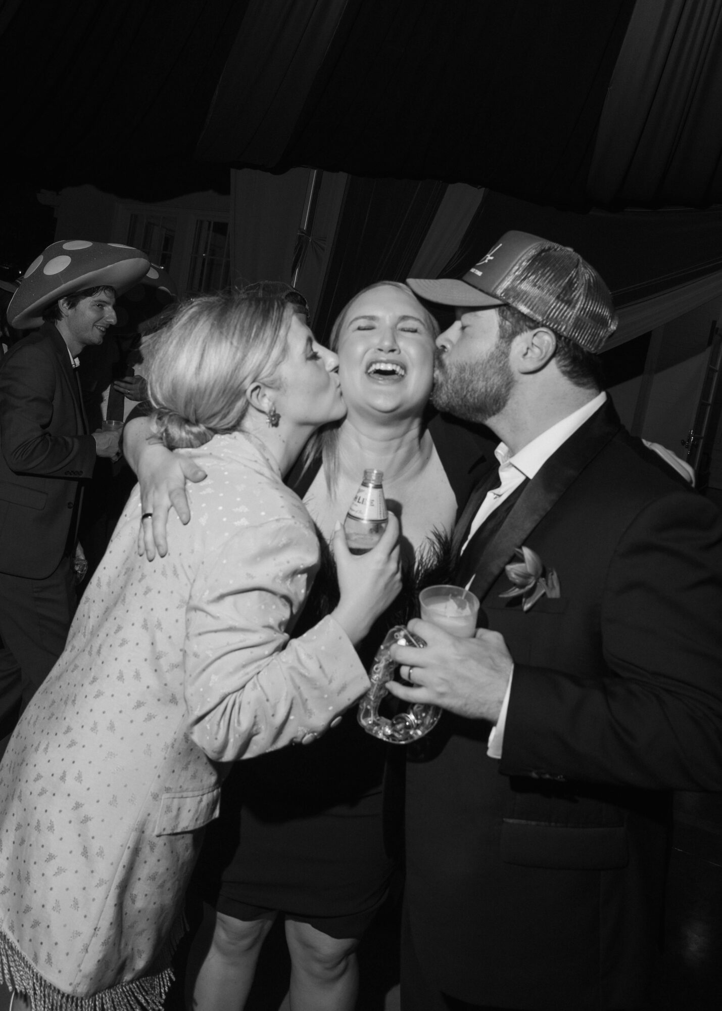 Three people laughing and kissing at a social event, with a woman in the center and two men on either side, one holding a drink.
