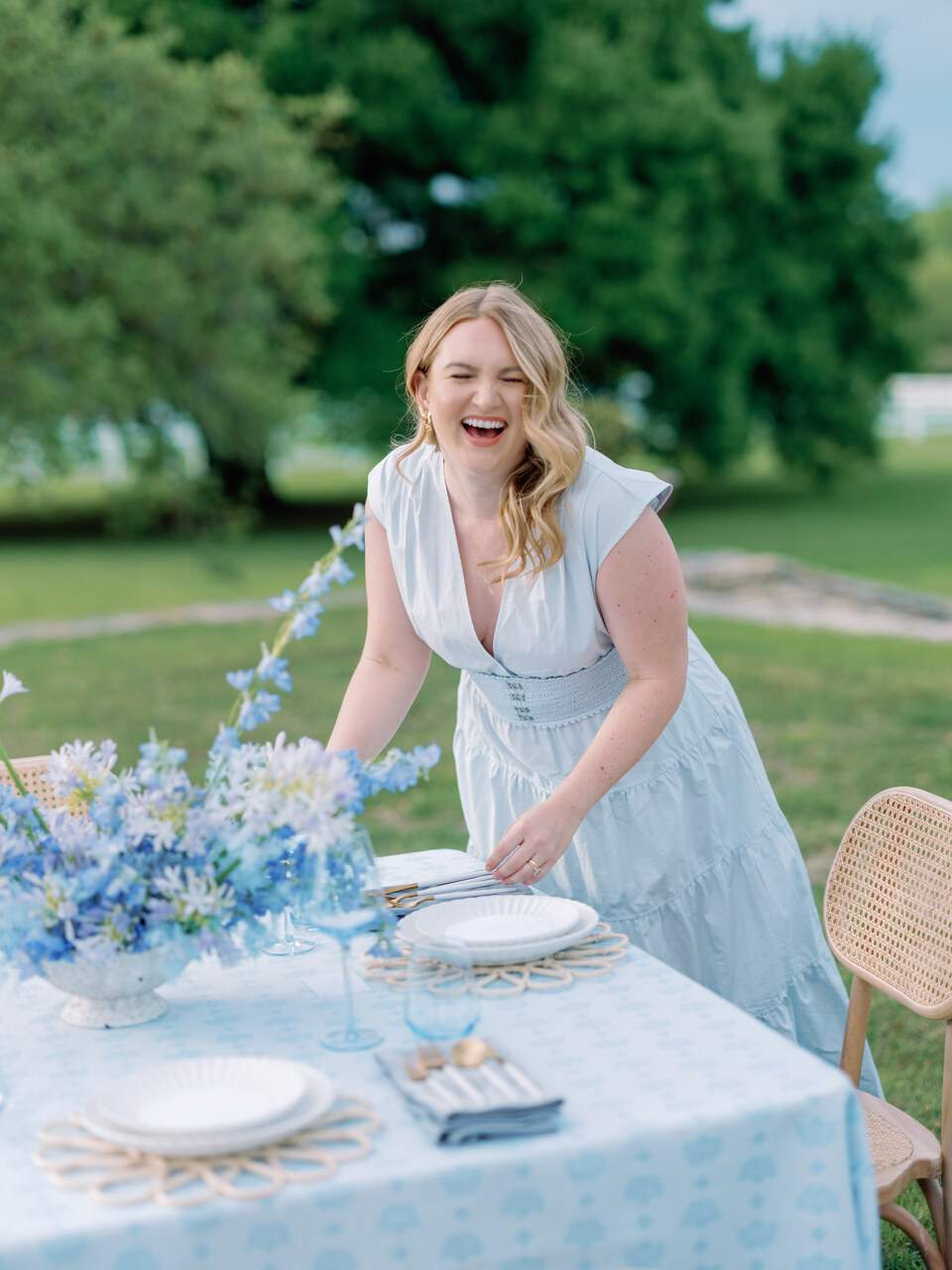 Woman in a light blue dress laughing outdoors at a table with flowers and tableware, greenery in background.