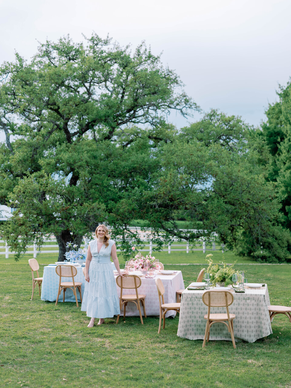 Two women in dresses stand near outdoor tables with chairs and tablecloths, set on a grassy area with trees and a fence in background.