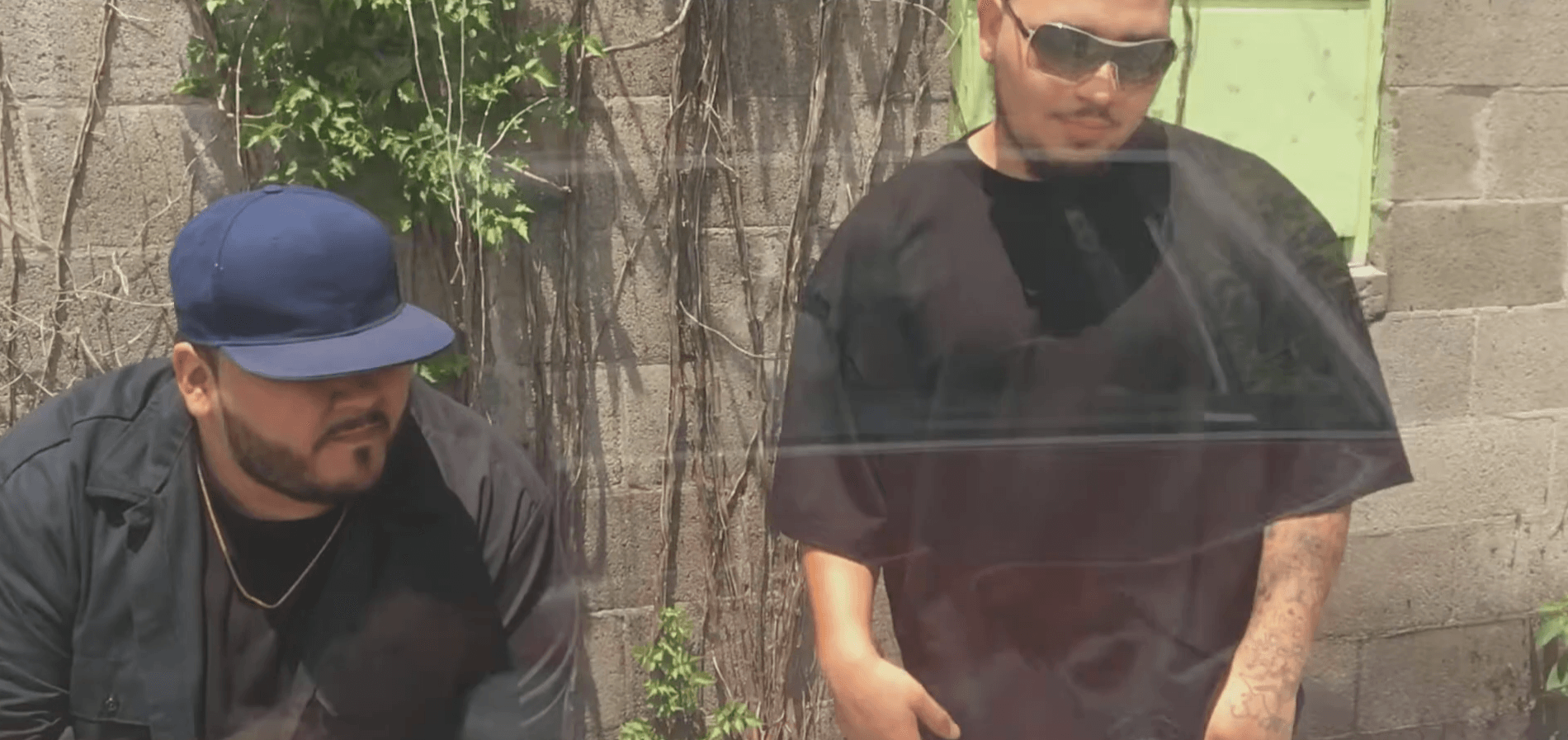 Two men standing outdoors near a wall and greenery, one wearing a cap and sunglasses, the other with sunglasses and a black shirt.