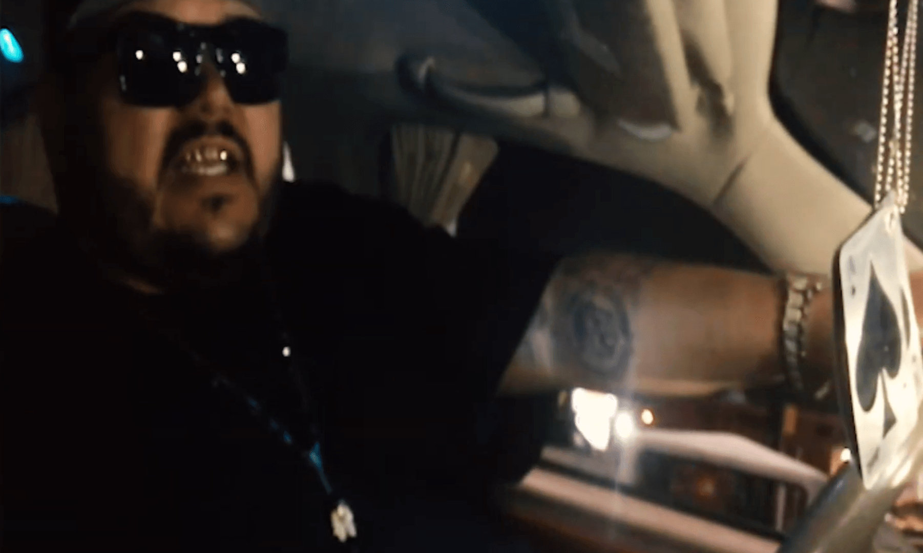 Person wearing sunglasses and a black shirt, sitting in a vehicle, with a tattoo visible on arm, and a chain necklace.