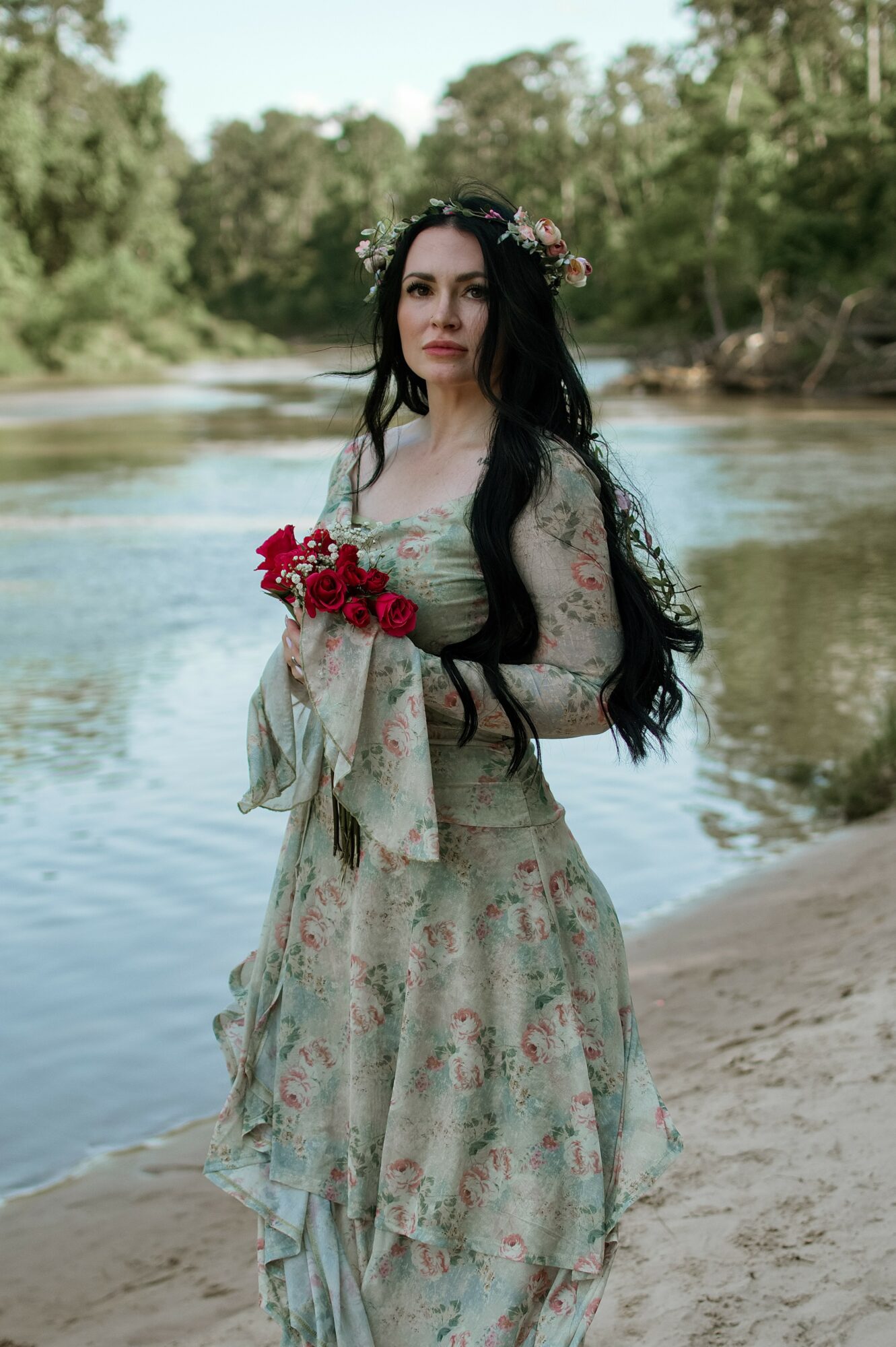 Woman in a floral dress holding red flowers by a river with trees in the background.