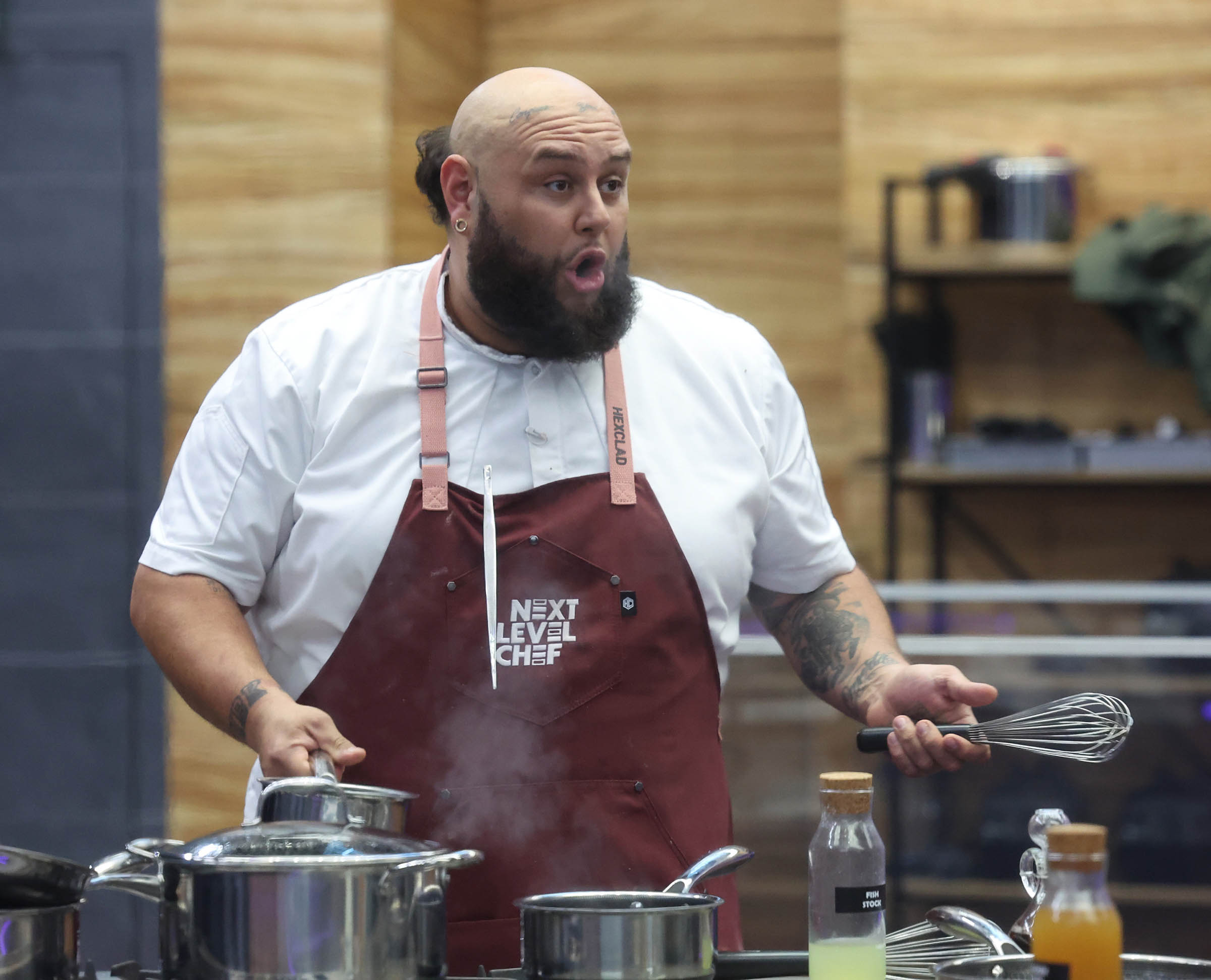 Man with a beard and bald head cooking, wearing a white shirt and maroon apron, in a kitchen setting.