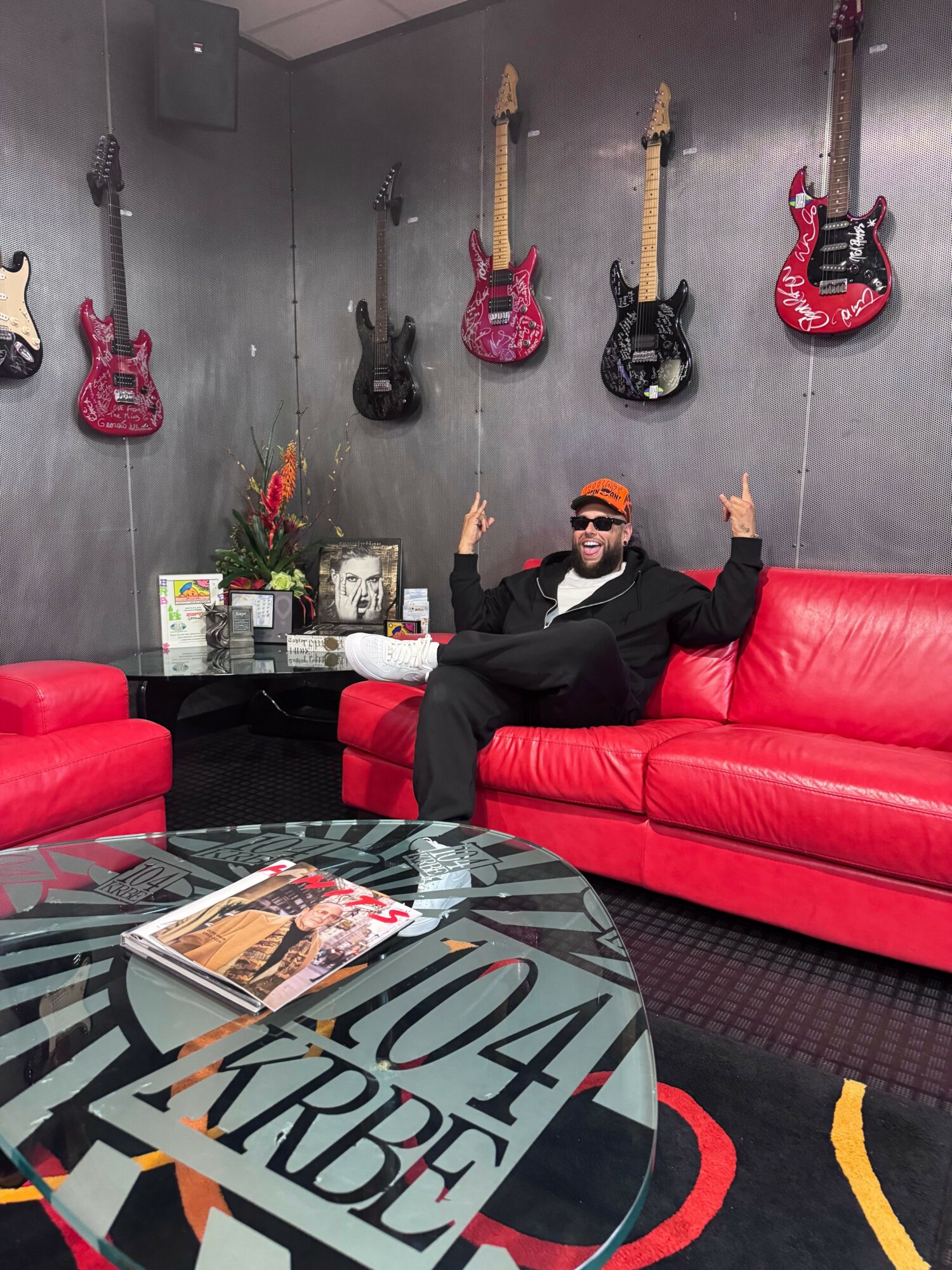 Person sitting on a red couch in a room with guitars hanging on the wall, holding a guitar, with a magazine on a table.