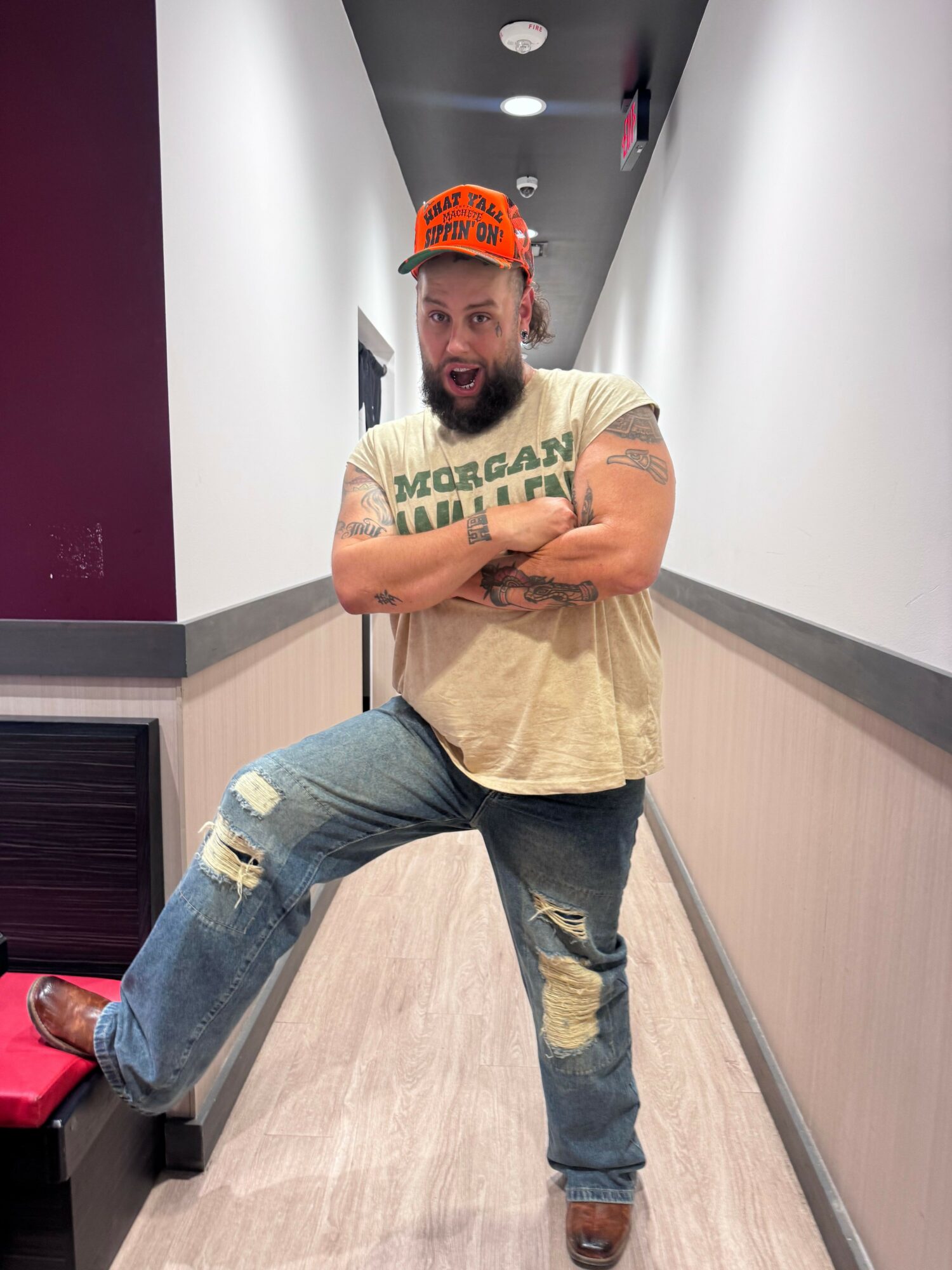Man standing in hallway, arms crossed, wearing a beige shirt, ripped jeans, and a red cap, looking at camera.