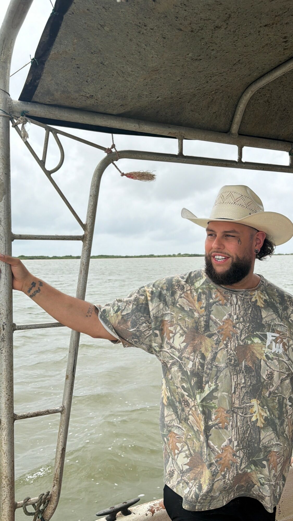 Man wearing a cowboy hat and camouflage shirt standing on a boat, holding a railing, with water and cloudy sky in background.