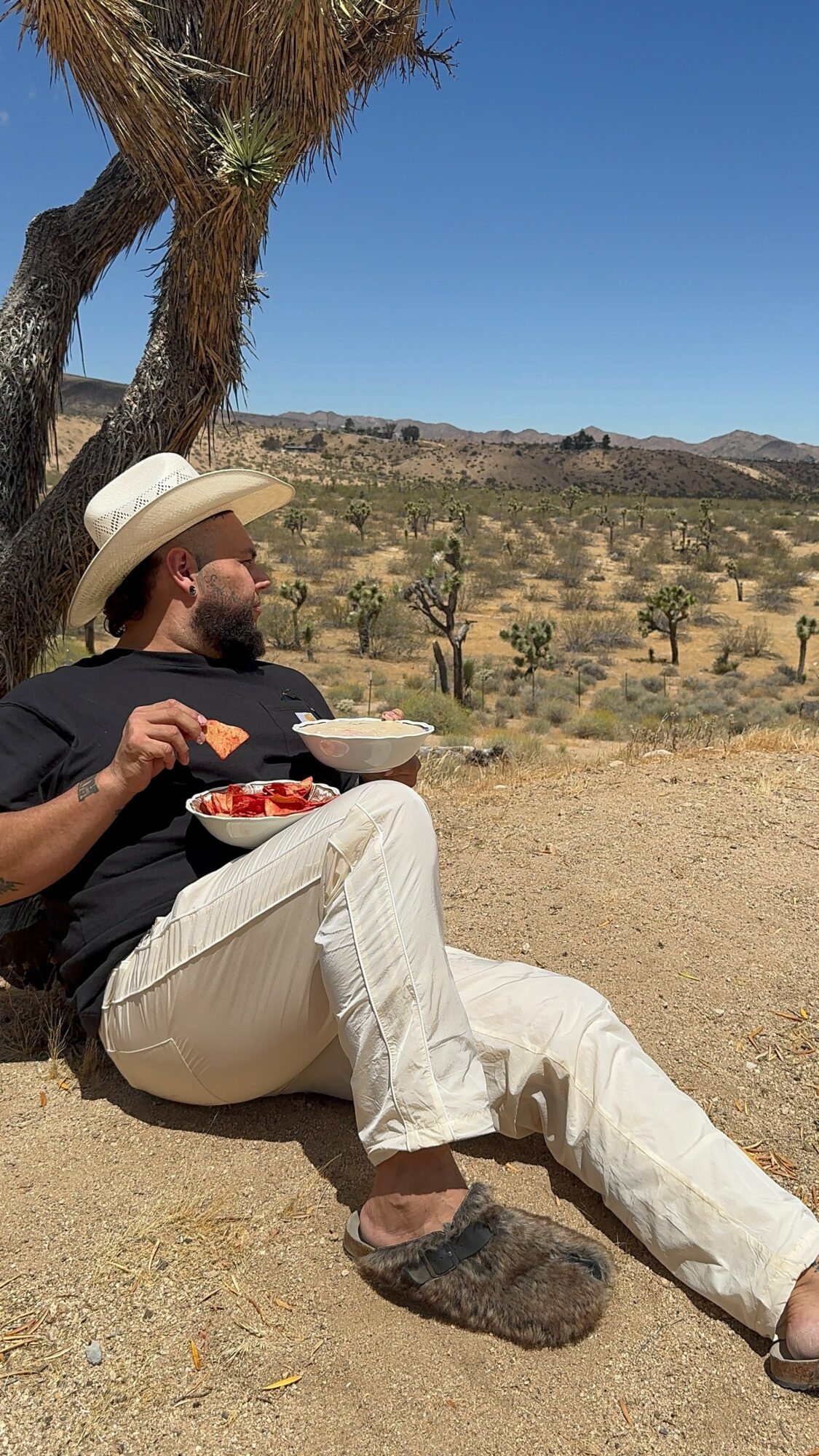 Person with a hat sitting against a tree, eating outdoors in a desert landscape with cacti and mountains.