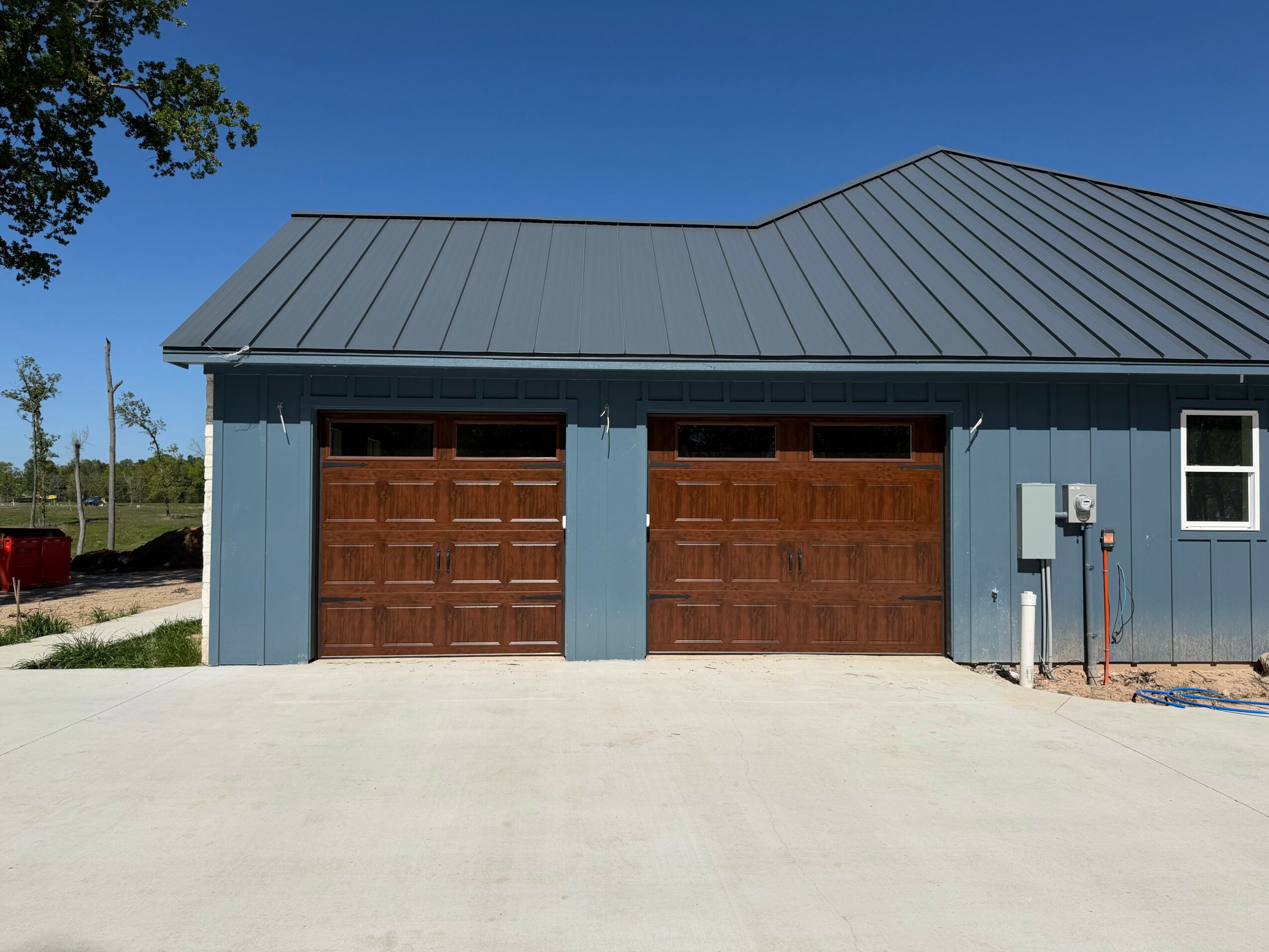 Blue building with two brown garage doors, metal roof, small window, electrical box, and clear sky.