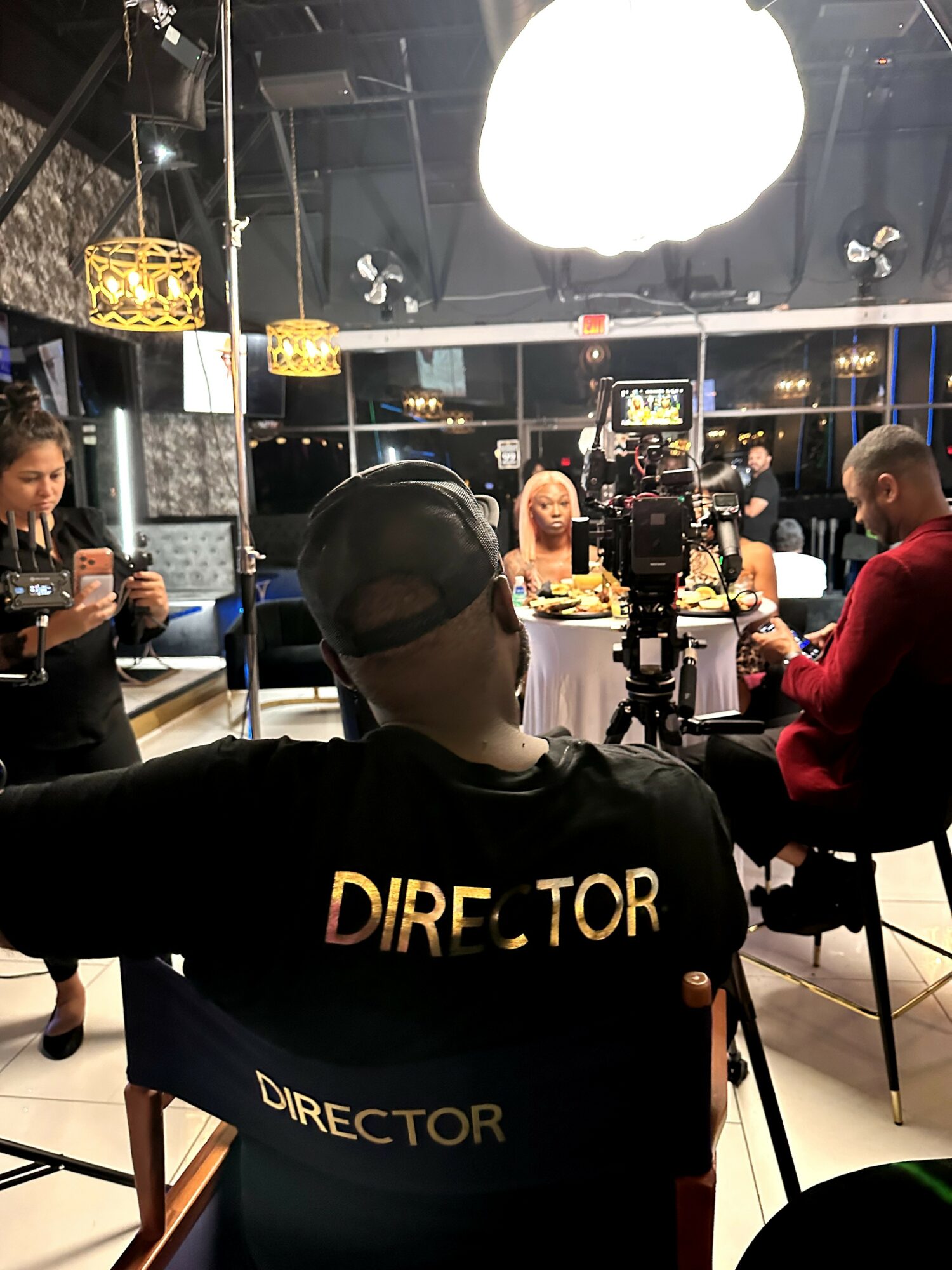 Person wearing a black shirt with 'DIRECTOR' written on the back, sitting at a table with people in a studio or set, bright lights overhead.