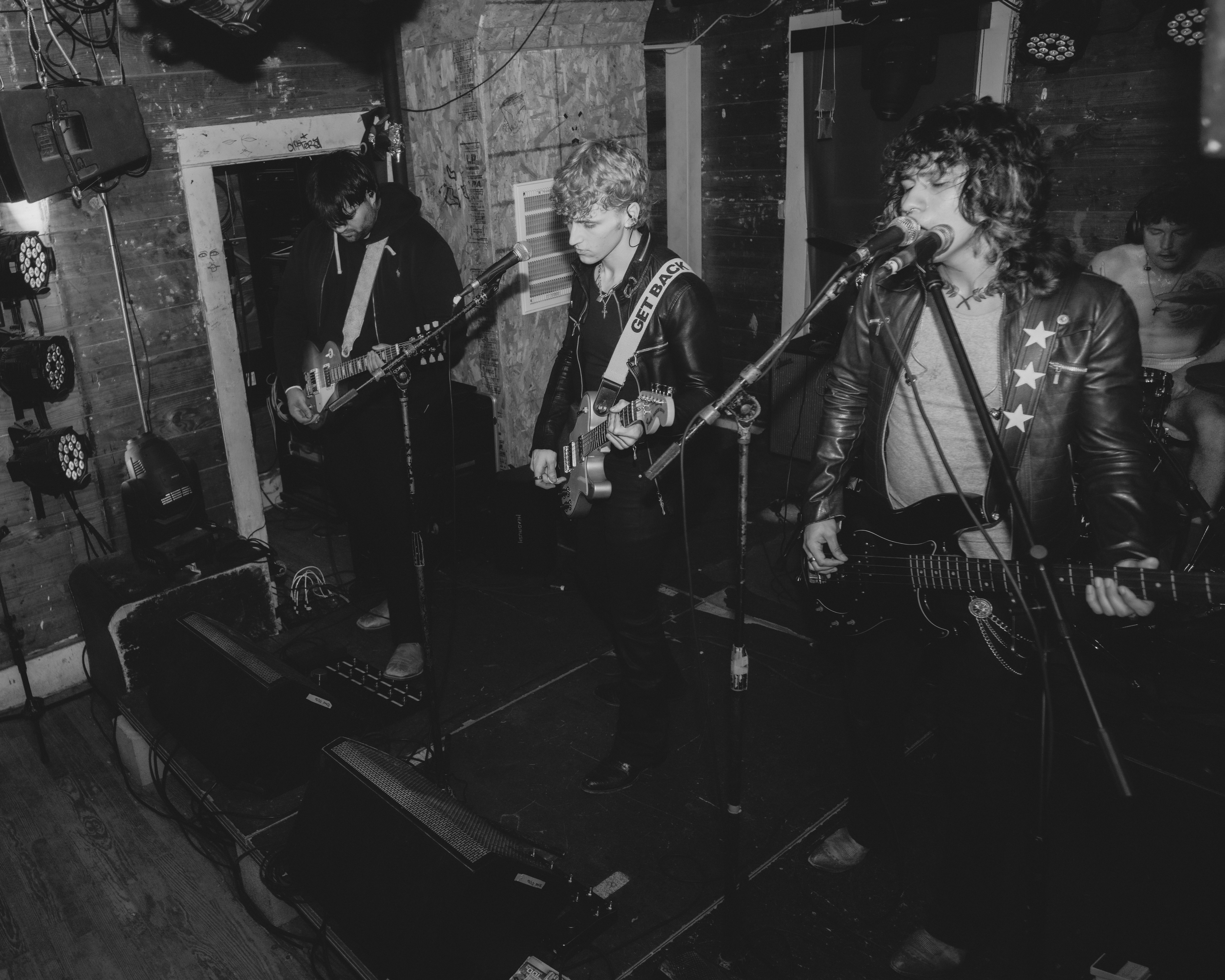 Three musicians perform on stage with guitars and microphone, in a dimly lit venue with brick walls.