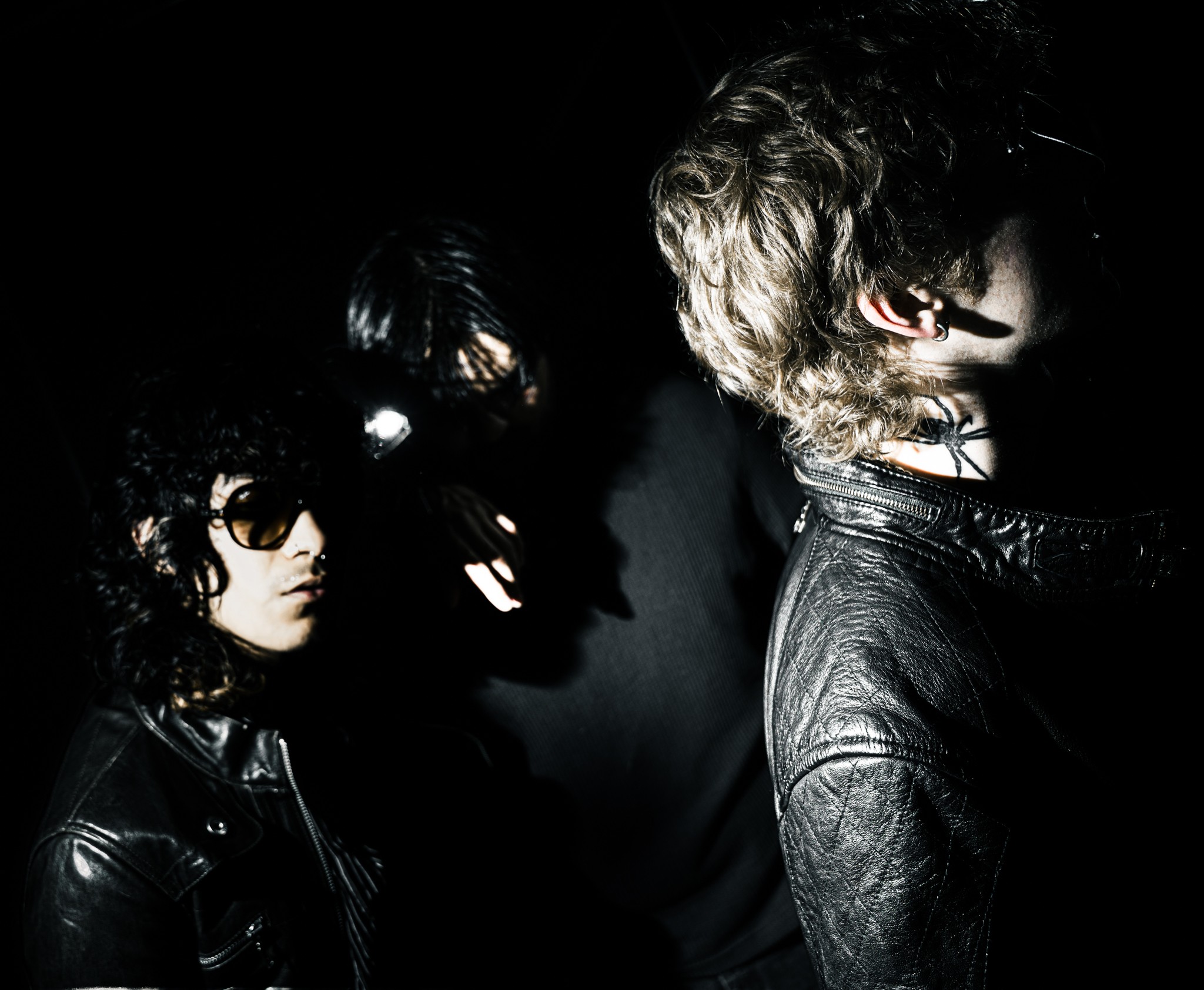 Three people in dark clothing and sunglasses, with one person wearing a mask, in a dark setting.