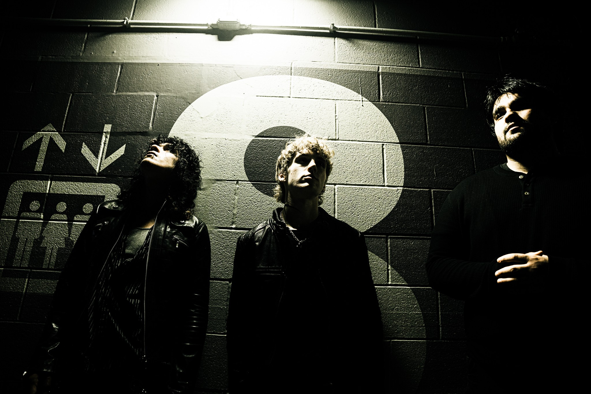 Three people standing against a wall with a target symbol, illuminated by a bright light above, dark atmosphere.