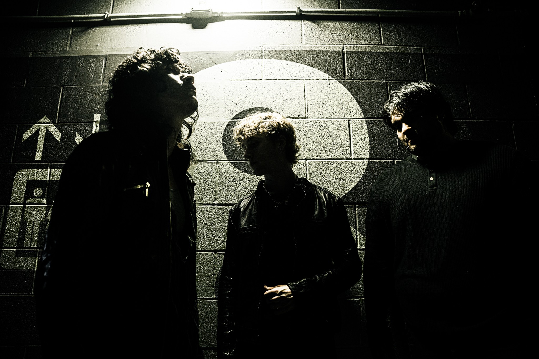 Three people standing in front of a wall with a target symbol, silhouetted against a light source, in black and white.
