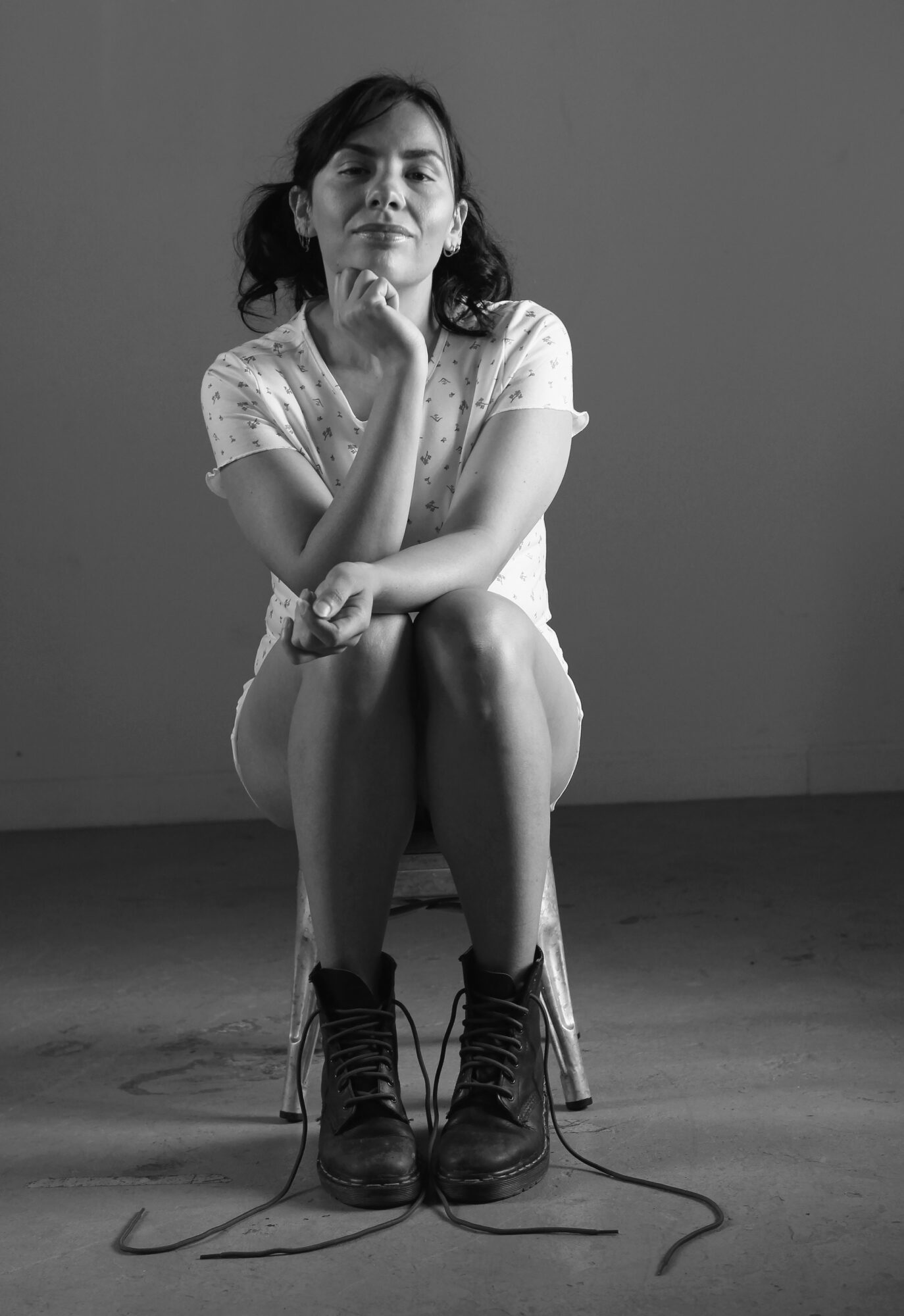 Young woman sitting on a chair with legs crossed, wearing a light-colored dress and boots, in a plain room.