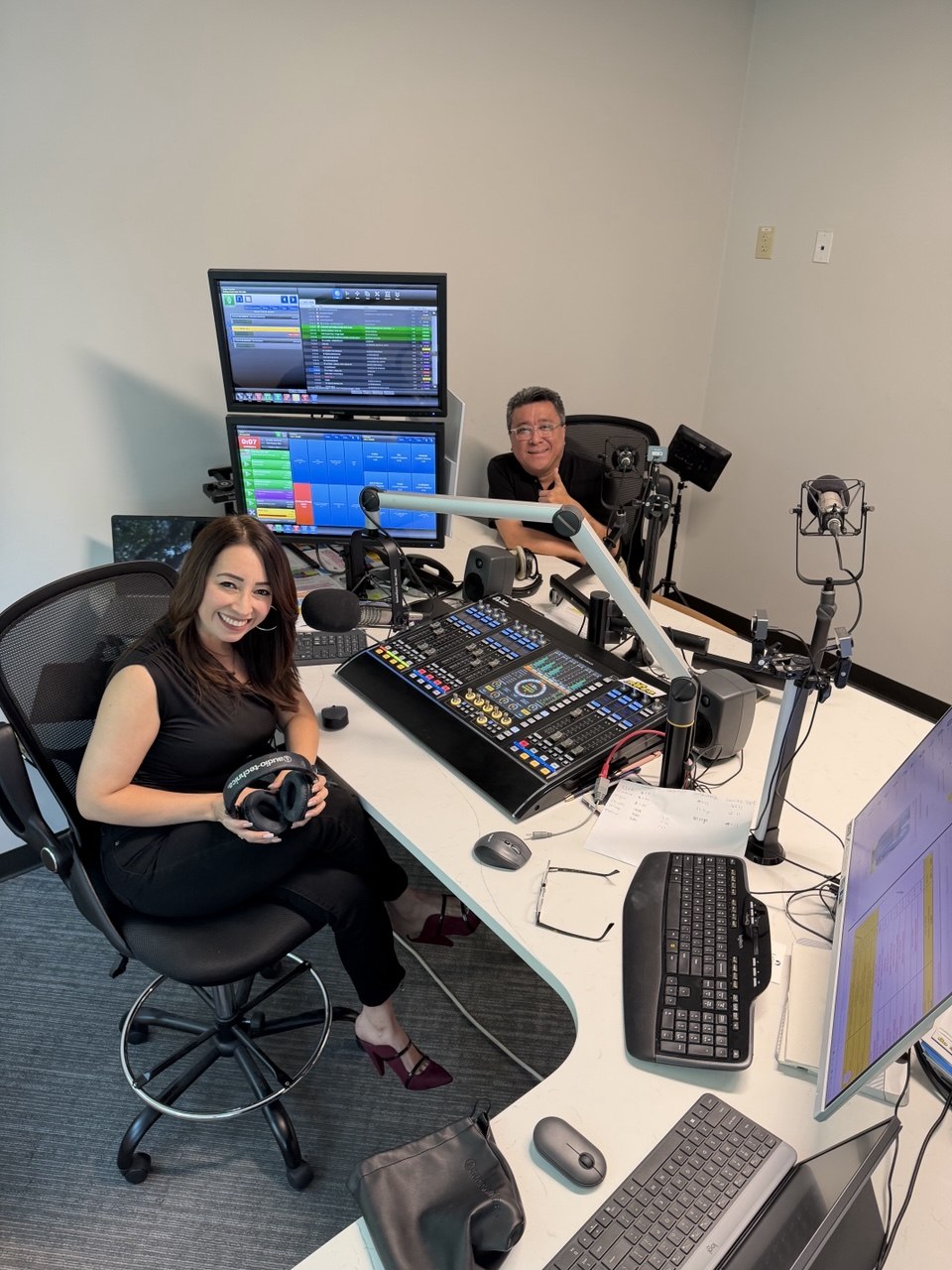 Two people in a radio studio with broadcasting equipment, microphones, and computer screens, smiling at the camera.