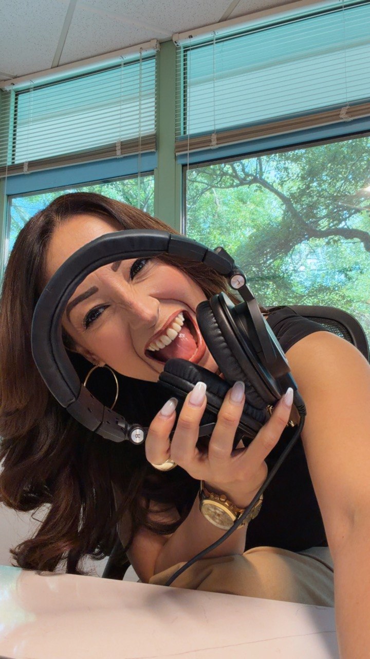 Woman holding headphones, smiling, indoors with large windows and trees outside, sunlight streaming in.
