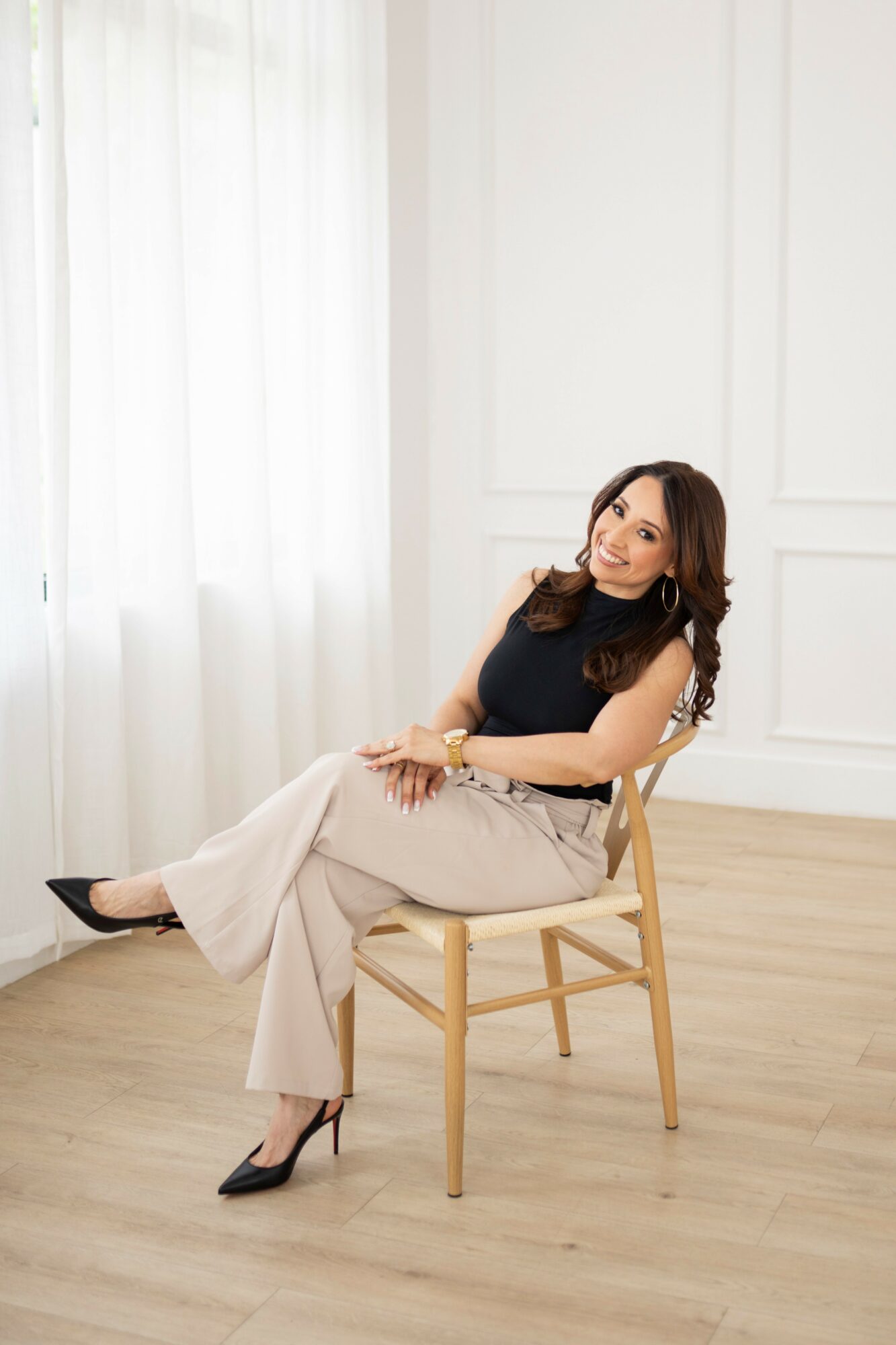 Woman sitting on a wooden chair in a bright room with white walls and curtains, wearing a black top and beige pants.