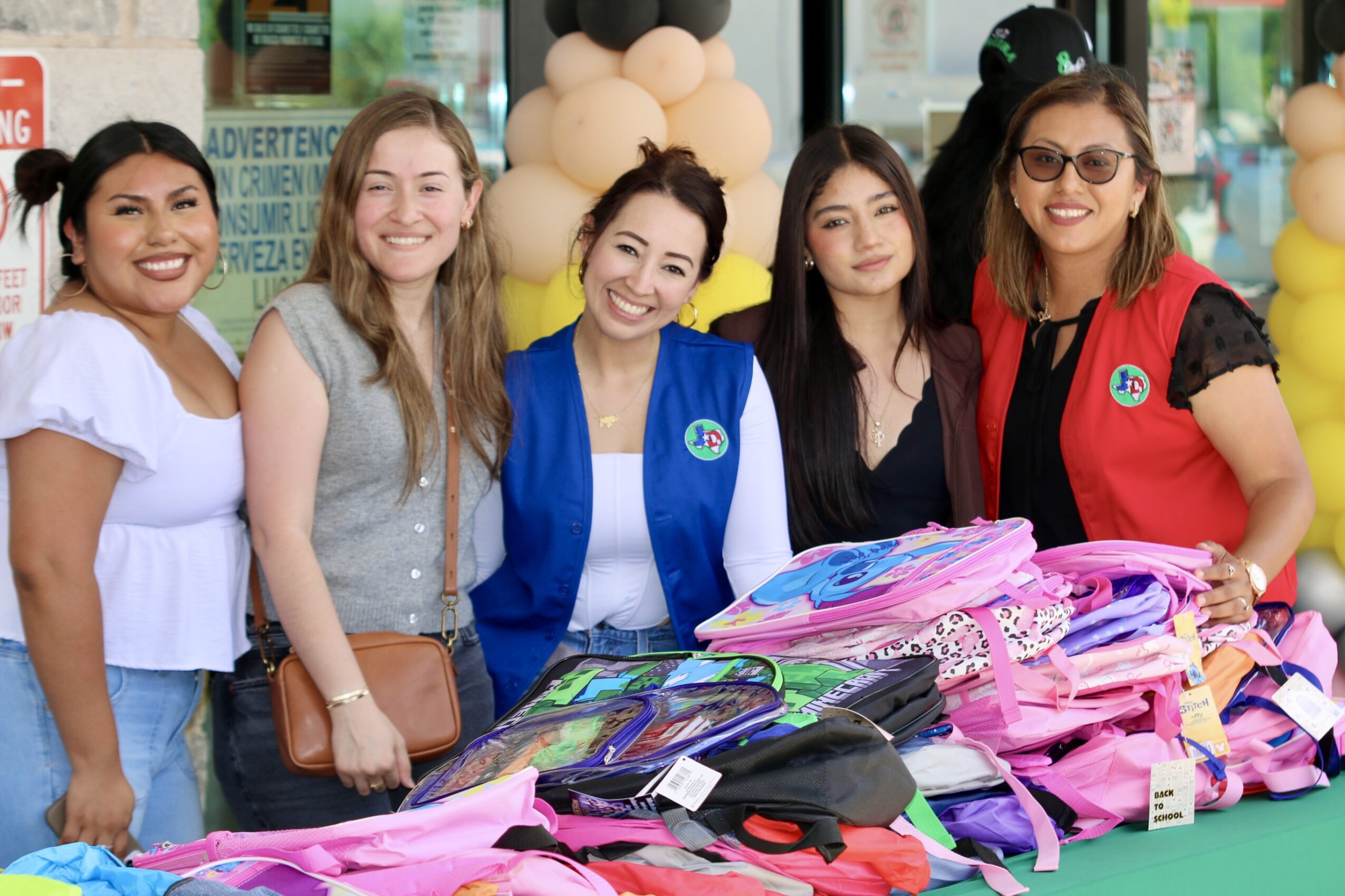 Five women standing behind a table with colorful bags and clothing, smiling at an outdoor event.