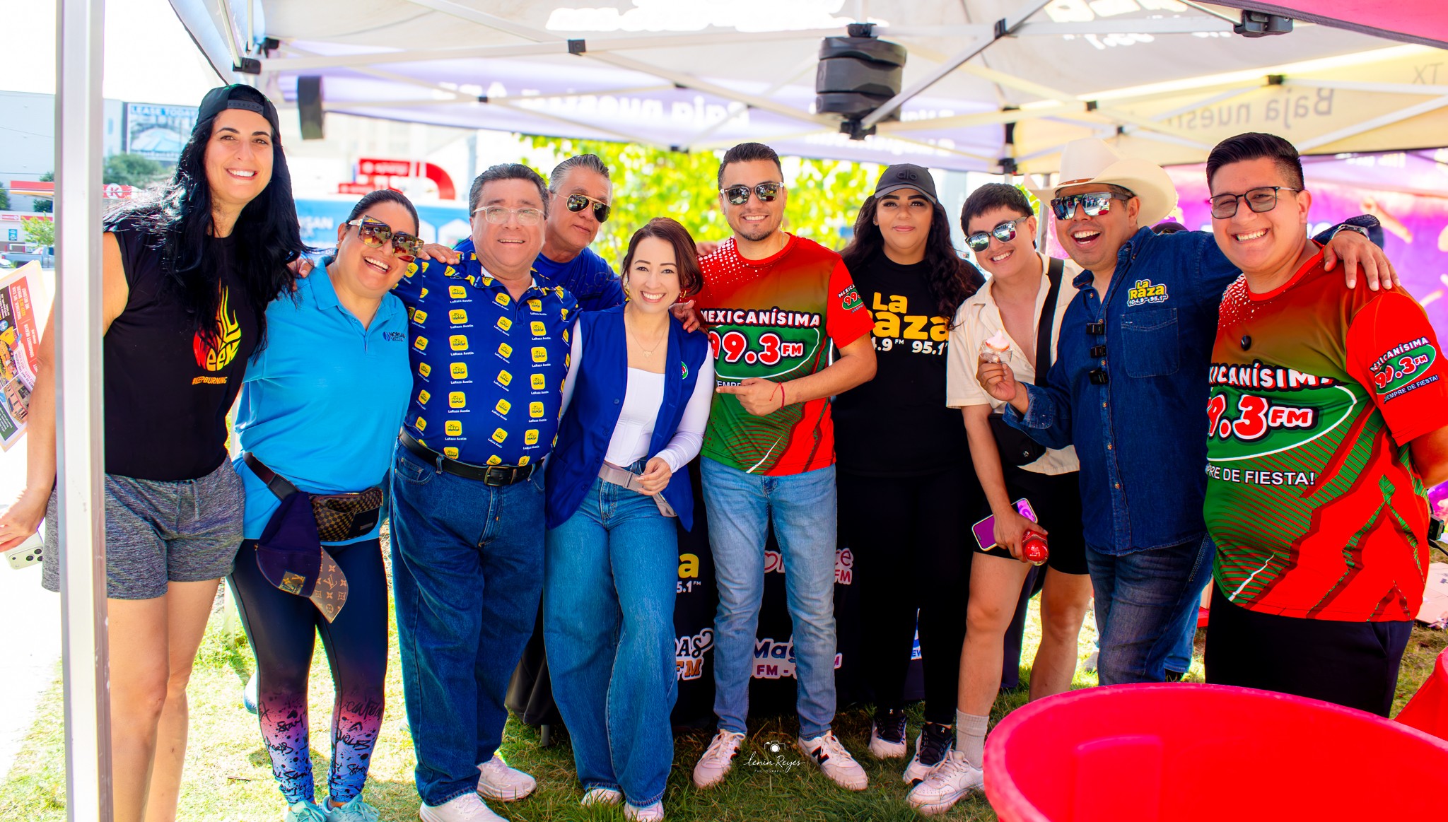 Group of ten people standing outdoors under a tent, smiling, with some wearing colorful shirts and sunglasses.