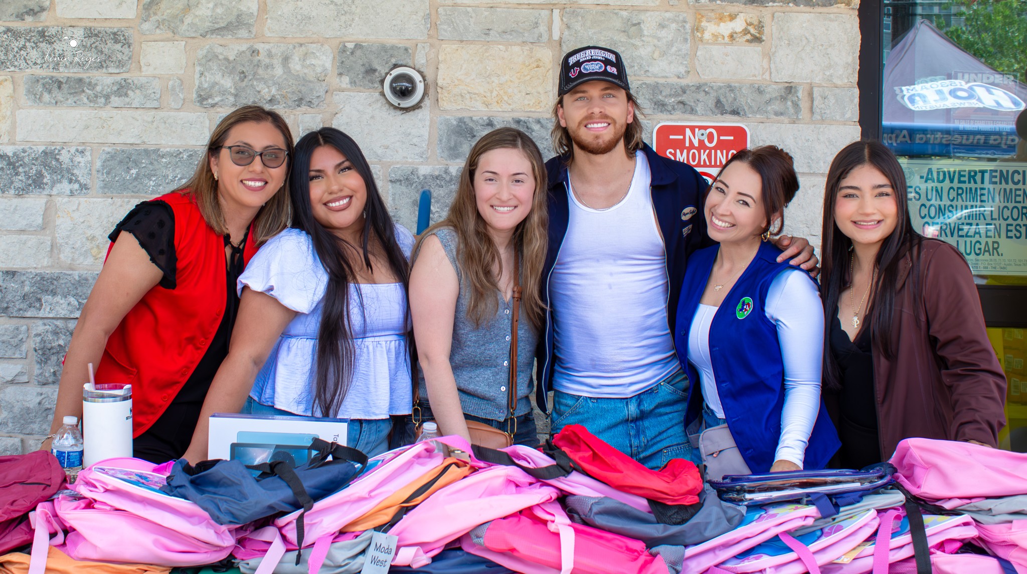 Group of six people standing behind a table with pink bags, smiling outdoors against a stone wall.