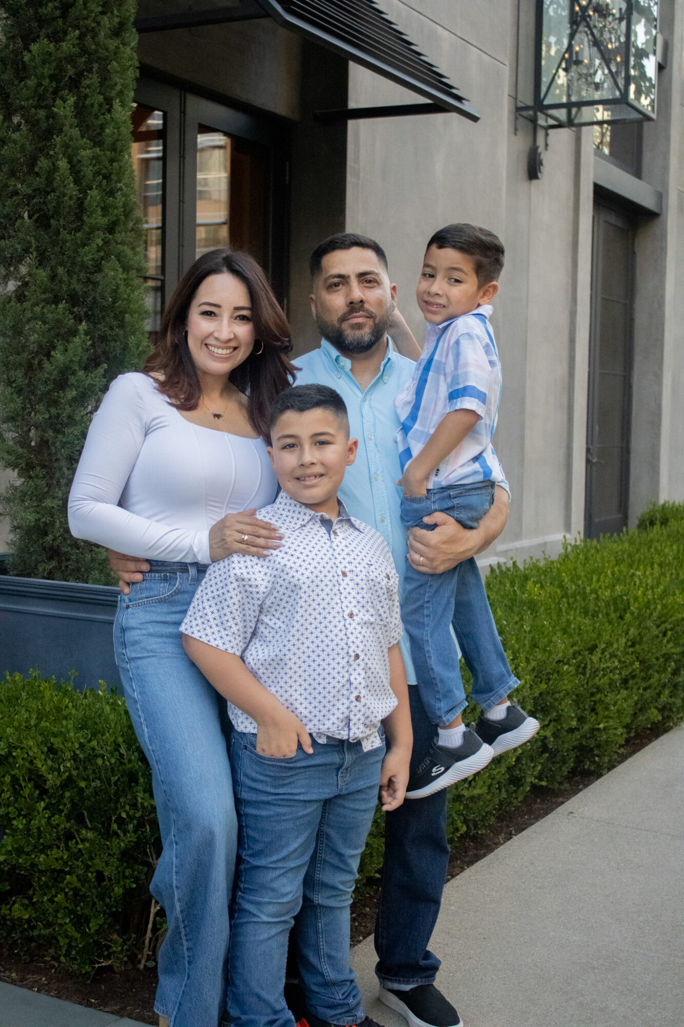 Family of four standing outdoors in front of a building, smiling, with greenery around them.