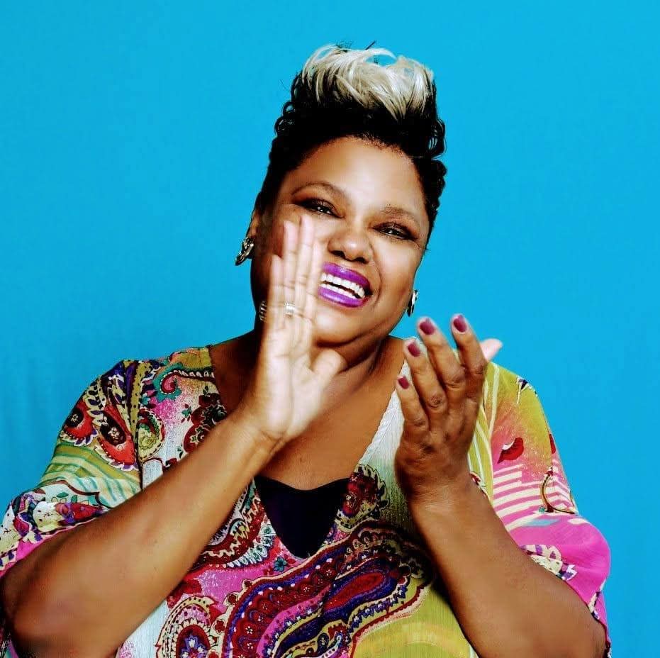 Woman with short hair and colorful patterned top, smiling and clapping hands, against a blue background.