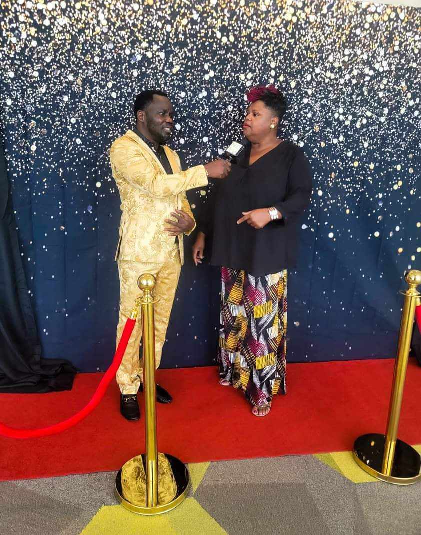 Two people stand on a red carpet with a glittery backdrop, one holding a microphone, engaged in conversation.